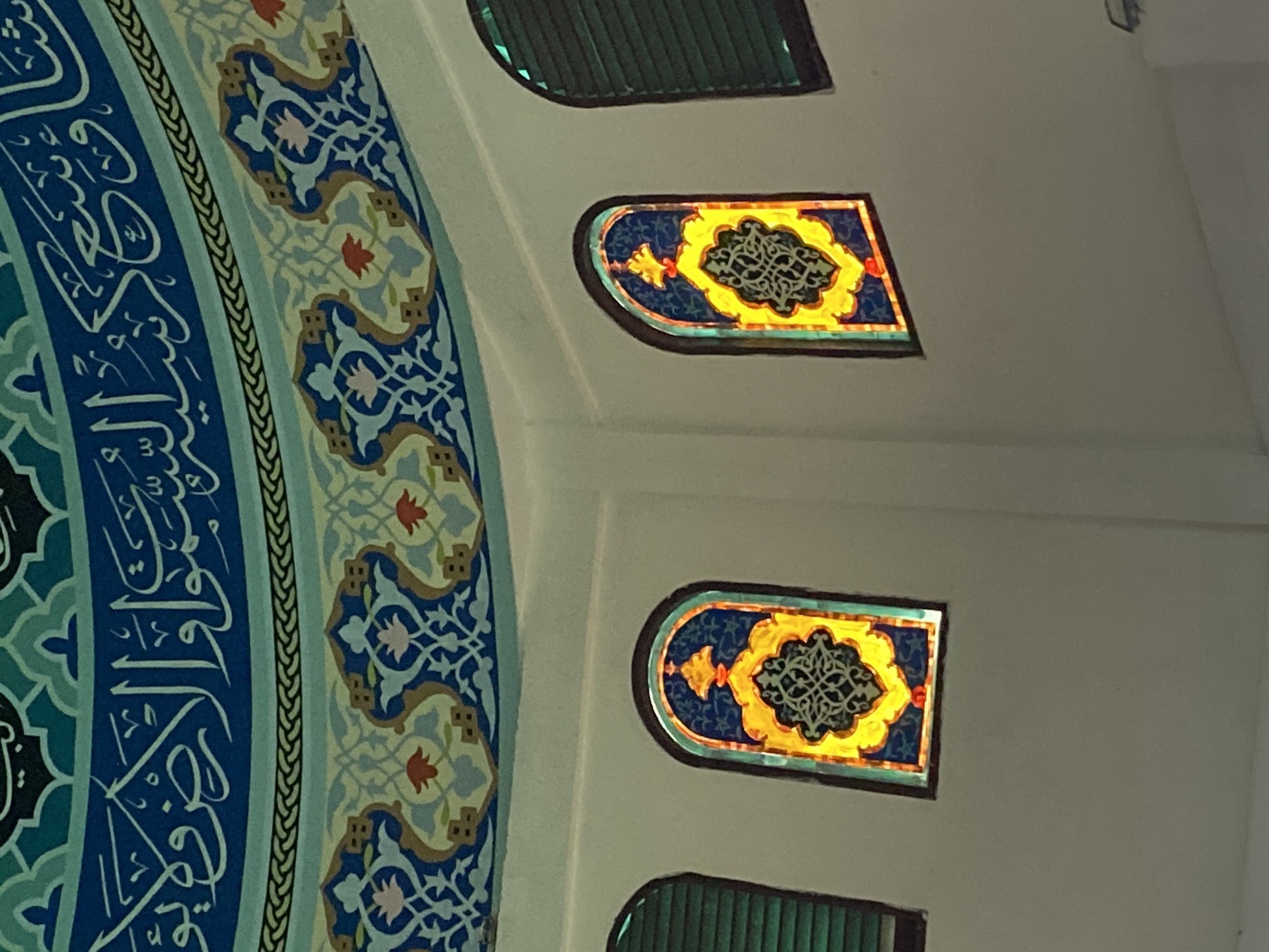 This image shows the interior of the Islamic Center Lhokseumawe, located in Lhokseumawe, Indonesia. The perspective is looking upwards towards the dome and walls of the building. The dome is decorated with intricate Islamic calligraphy and geometric patterns in shades of blue, green, and yellow. Below the dome, on the curved white walls, are several arched windows. Two of these windows are visible in detail and feature stained glass with ornate, symmetrical designs in vibrant yellow, blue, and red hues. The windows on either side of these stained glass panes appear to be covered with horizontal green blinds. The overall impression is one of rich ornamentation and religious architecture.