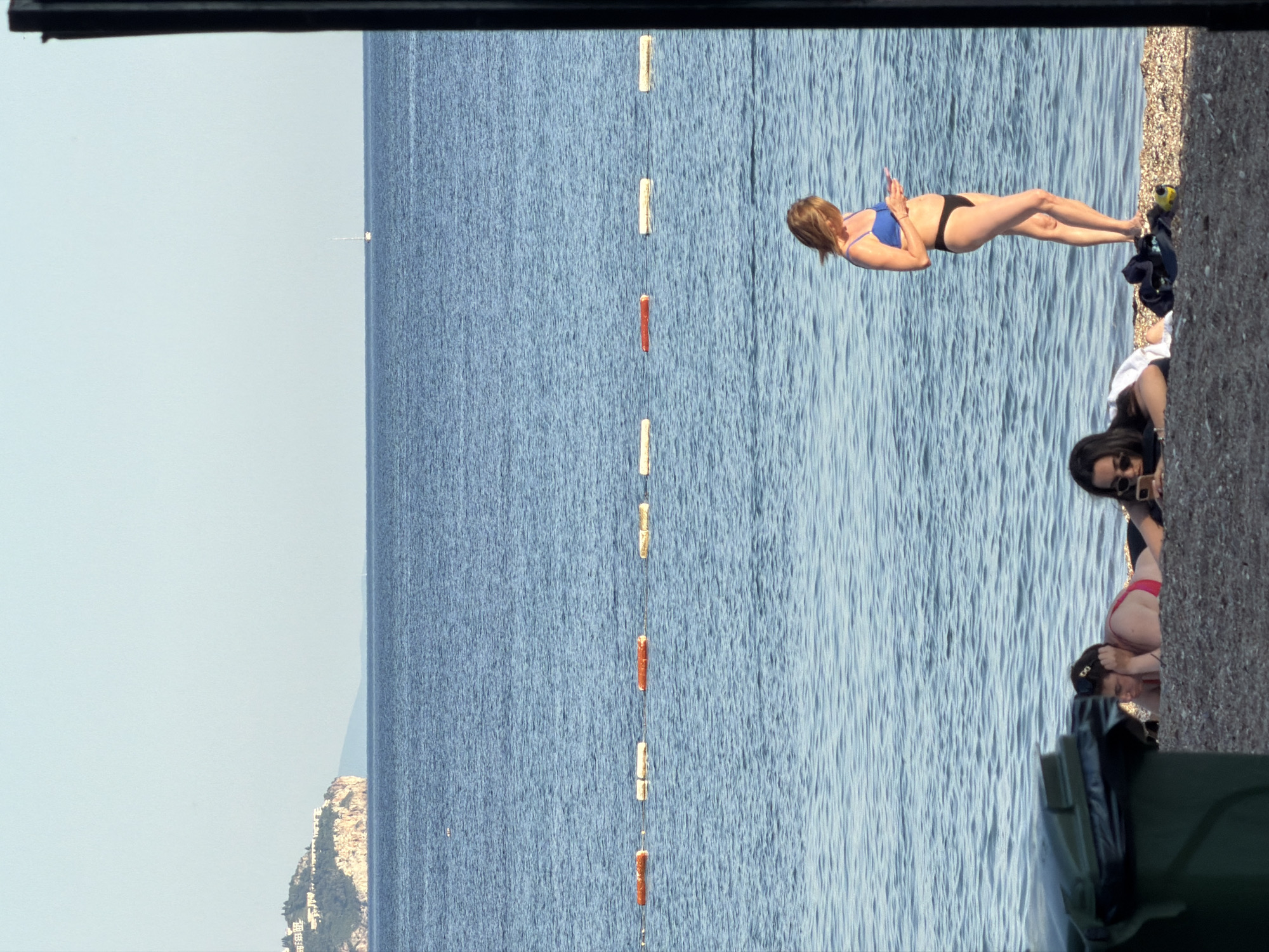 The image captures a beach scene in Budva, Montenegro. The view is oriented vertically, with the sky and distant mountains visible to the left, the Adriatic Sea occupying the majority of the central area, and a beach area to the right. Buoys are visible in the sea, possibly marking a designated swimming area. A woman in a blue bikini is lying on the beach, facing the sea and appearing to use a mobile device. Nearby, two other women are present: one with sunglasses is using her phone. A trash can is visible in the foreground. The environment suggests a sunny day at a popular beach location.