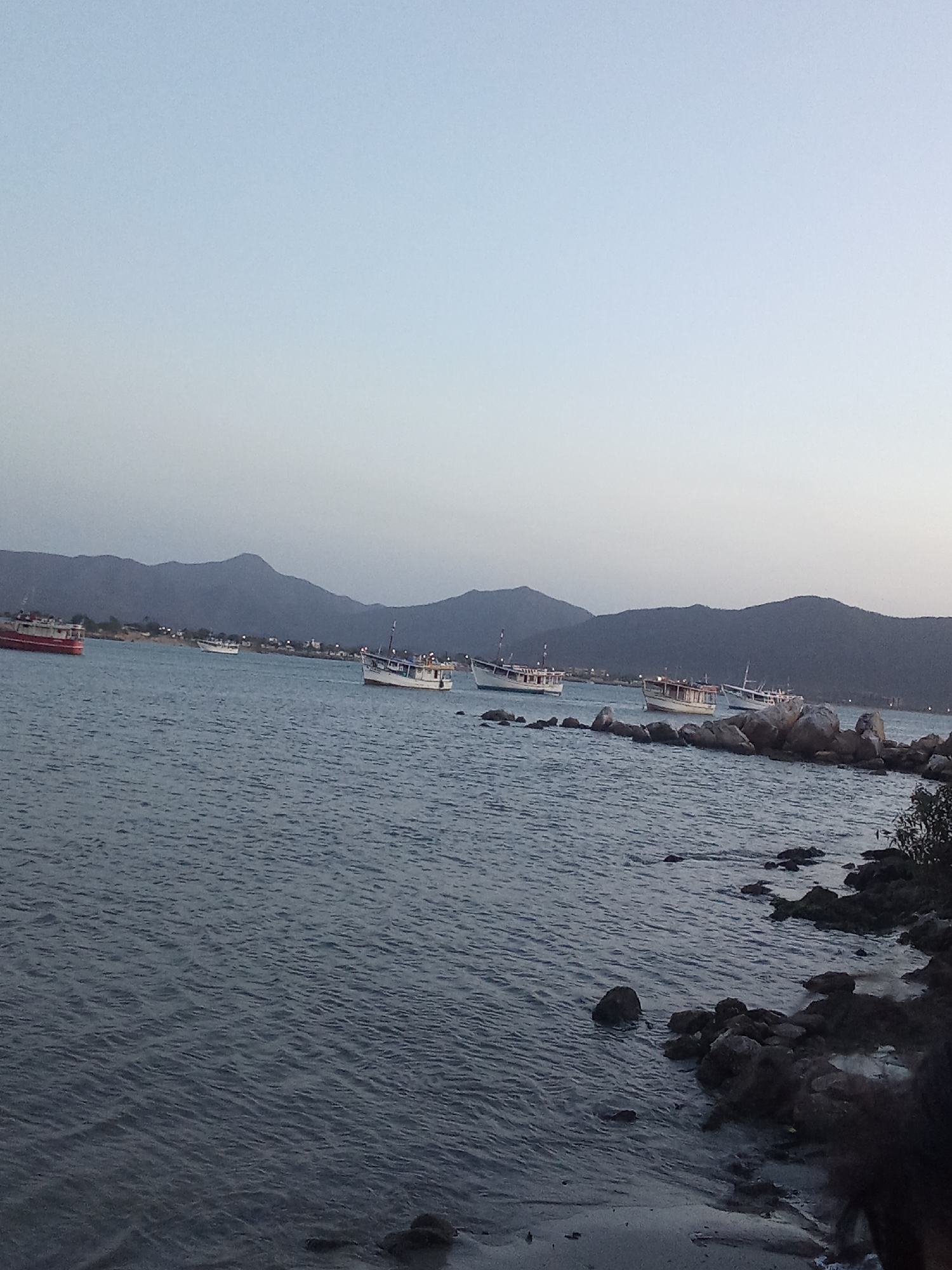 The image depicts a coastal scene in Juan Griego, Venezuela. Several small fishing boats are anchored in a bay. Low mountains are visible in the background under a pale sky. The foreground shows a rocky shoreline with a dark sandy beach. The water appears calm, and the lighting suggests either dawn or dusk. There are no visible people.