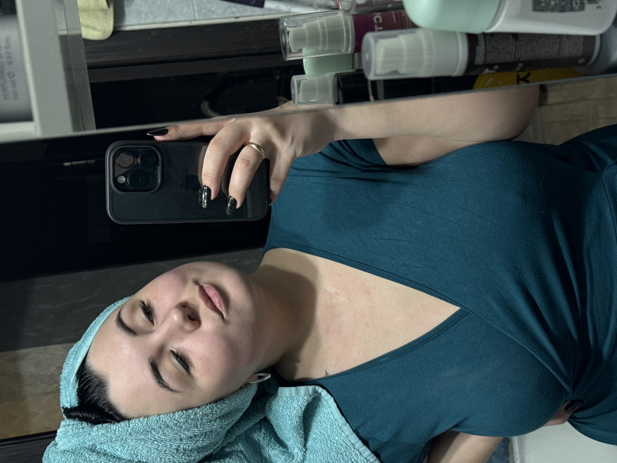 A woman is taking a selfie in what appears to be a bathroom. She is wearing a teal-colored top and has a light blue towel wrapped around her head. She is holding a black smartphone in her right hand, and the reflection in the mirror shows bottles of skincare products on a shelf behind her. The background and the overall composition suggest a domestic, personal setting. The location is unknown.