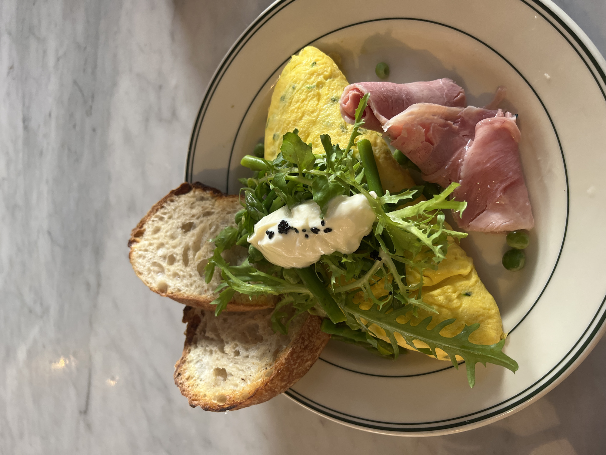 A plate of food on a marble table. The plate has a green and white rim and contains an omelet, ham, salad, and bread. The salad is topped with a dollop of white cream and black seeds. Two slices of bread are on the left side of the plate. Some asparagus and peas are also on the plate.