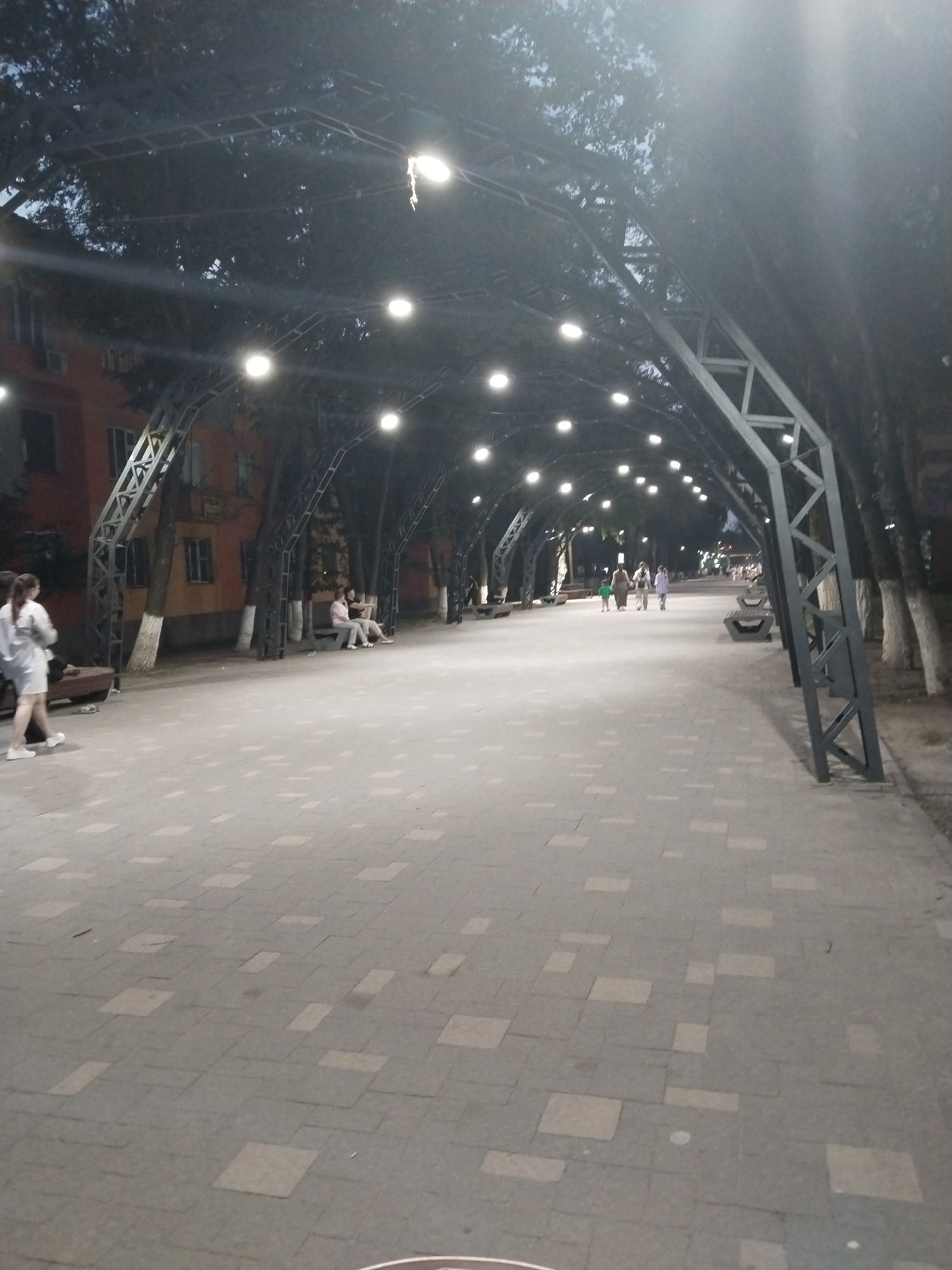 This image captures an outdoor scene at dusk, likely in the city of Ötegen batyr, Kazakhstan, focusing on a pedestrian walkway lined with trees and an overhead structure adorned with lights. The walkway is paved with grey tiles, some of which are lighter in color, creating a patterned effect. Along the sides of the walkway, there are benches and trees. An arched metal structure with a geometric design spans overhead, from which numerous round lights are suspended, illuminating the pathway. In the background, a building with orange walls and dark windows is visible, suggesting an urban or semi-urban setting.
Several individuals are present in the scene. On the left side of the walkway, a person wearing a light-colored dress and white shoes is walking away from the camera. Further down the walkway, on the left, two people are seated on a bench. Towards the middle ground, a group of three people, two adults and a child, are walking together. In the distance, more people can be seen strolling along the path, indicating a public space that is being utilized by residents. The overall atmosphere is calm and the lighting suggests the transition from day to night.