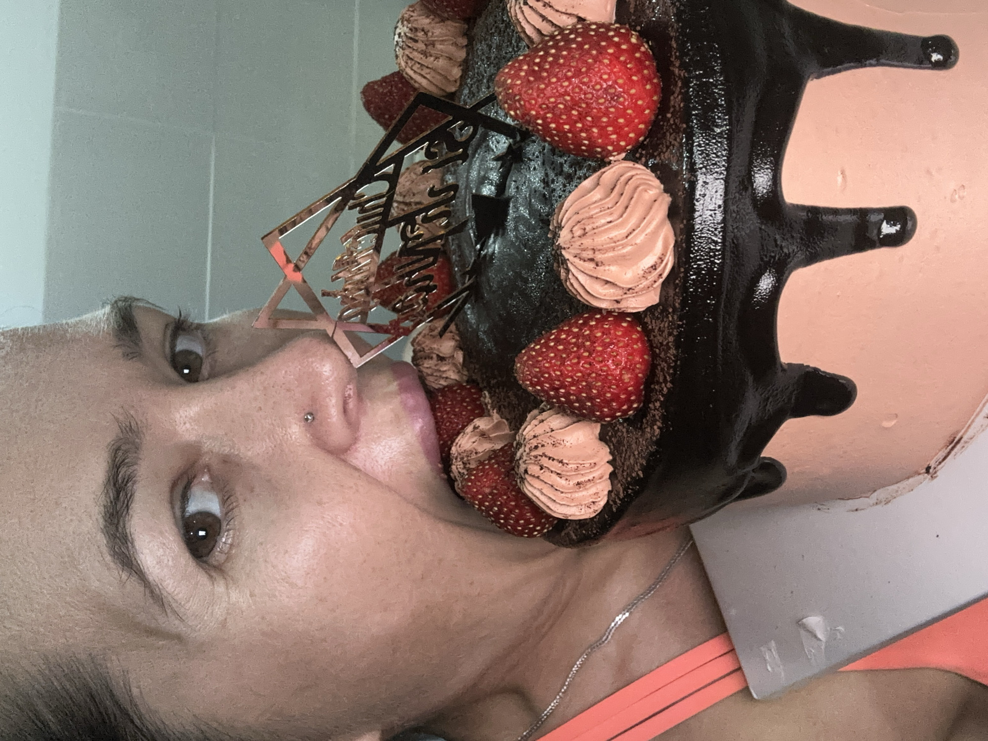 A rotated close-up image shows a woman's face positioned near a chocolate cake. The cake is decorated with strawberries and piped rosettes of a tan-colored frosting. A gold-colored cake topper is also visible. The woman has brown eyes and a nose piercing. Her lips are slightly parted. She is wearing a light orange top and a silver chain necklace. The background appears to be a wall, possibly tiled. There is no verifiable information to confirm the location of the image as Bac Nha Trang Ward, Vietnam.