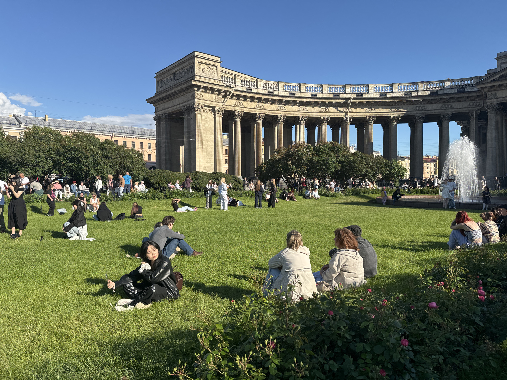 The image depicts a sunny day at Kazan Cathedral in Saint Petersburg, Russia. A large semicircular colonnade of the cathedral serves as a backdrop to a green lawn where numerous people are scattered. Several individuals are sitting or lying on the grass, some in small groups, while others are alone. In the foreground, a woman in a black leather jacket is taking a selfie with her phone while sitting cross-legged on the grass. In the background, a fountain is visible, and more people are gathered near it. Trees and bushes are interspersed across the lawn, adding to the park-like atmosphere. The sky is clear and blue, with a few wispy clouds. The scene conveys a sense of leisure and public gathering on a pleasant day.