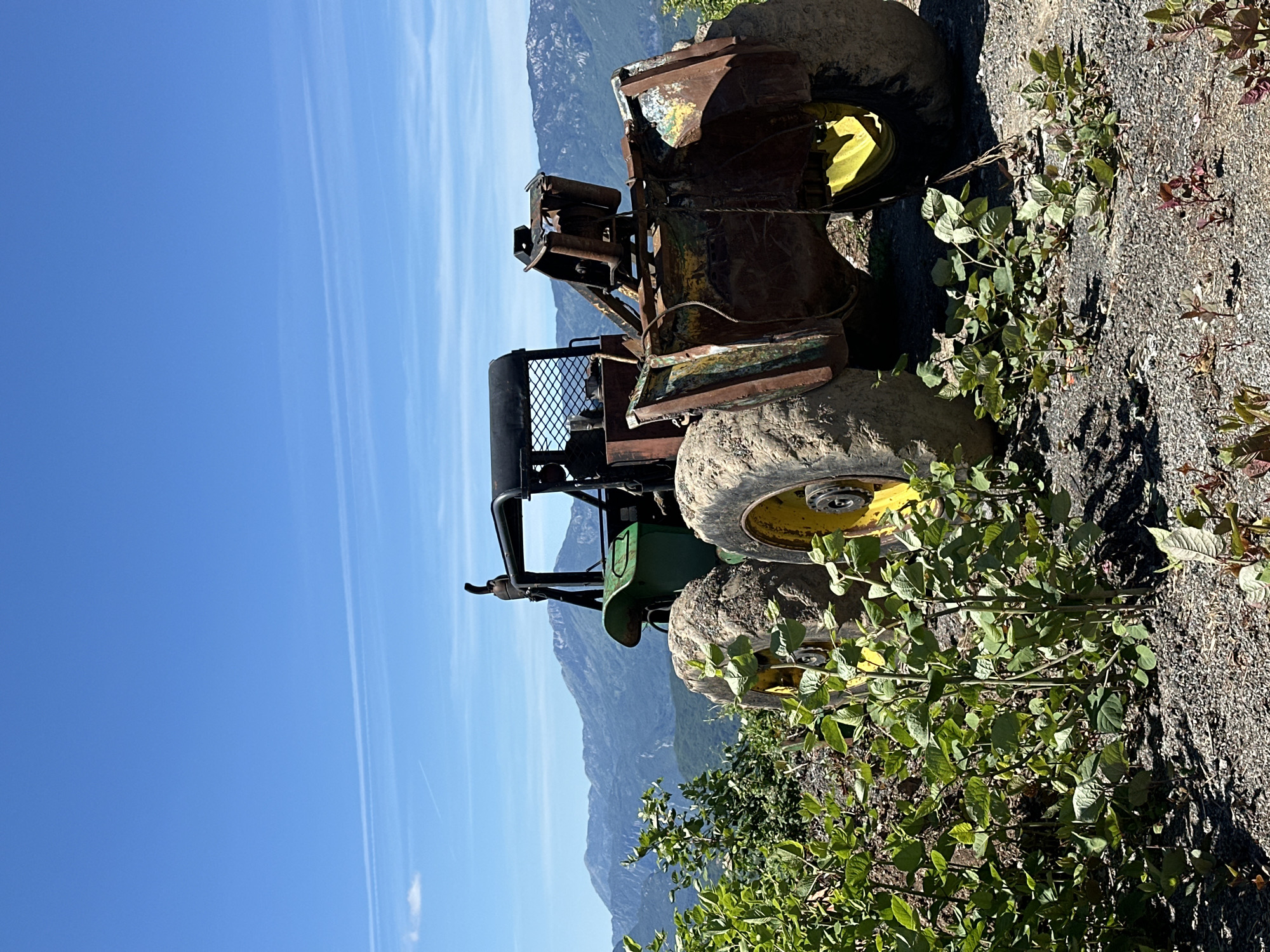 An abandoned, rusty, and weathered farm tractor sits on a gravelly, mountainous terrain in Crkvine, Montenegro. The tractor, predominantly green and brown with yellow accents on its wheels, is positioned at an angle, facing right. Its large, mud-caked tires are prominent. Vegetation, consisting of leafy green plants and shrubs, grows around the tractor and in the foreground, some of which appears to be encroaching on the machinery. In the background, a vast mountain range stretches across the horizon under a clear blue sky with a few wispy clouds and contrails. The overall scene suggests a remote, possibly disused, location within a natural, mountainous landscape.