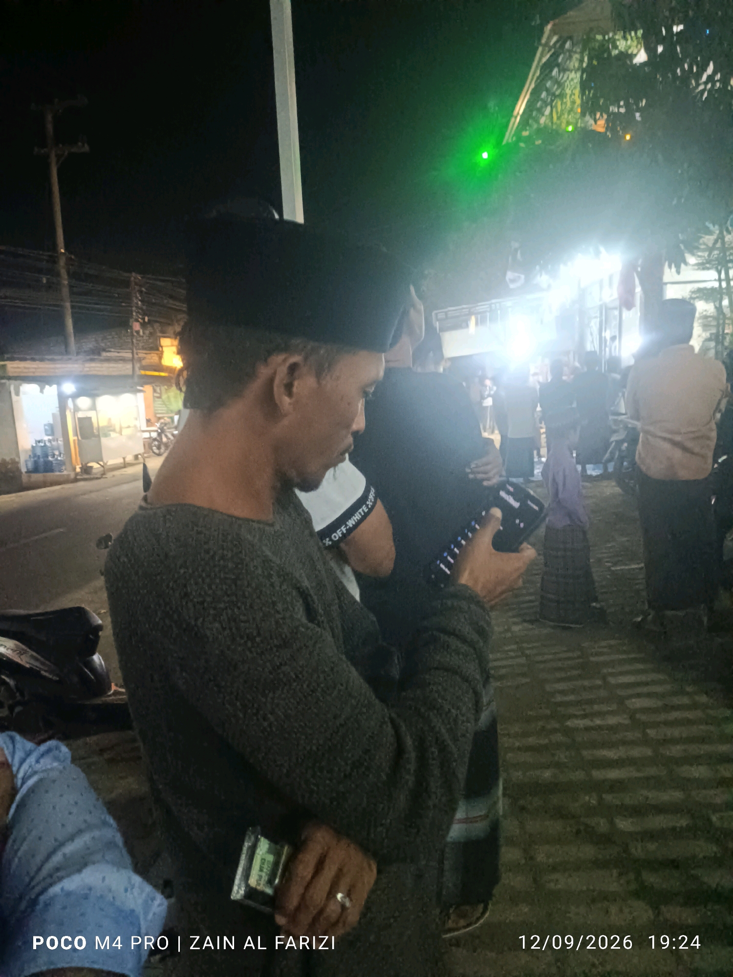 The image shows a night scene outdoors, possibly in a public area of Cendagah, Indonesia. A man in the foreground, wearing a dark cap and a grey sweater, is looking down at his phone, which he holds in his right hand. His left hand, on which he wears a ring, is visible near his hip. He is standing next to a motorcycle. In the background, several other people are visible, some wearing traditional Indonesian clothing such as sarongs. There are bright lights illuminating the scene, some of which appear to be from a stage or a gathering. A green light is visible in the upper right portion of the image. The ground appears to be paved with interlocking bricks. The time stamp "12/09/2026 19:24" and the text "POCO M4 PRO | ZAIN AL FARIZI" are superimposed on the image.