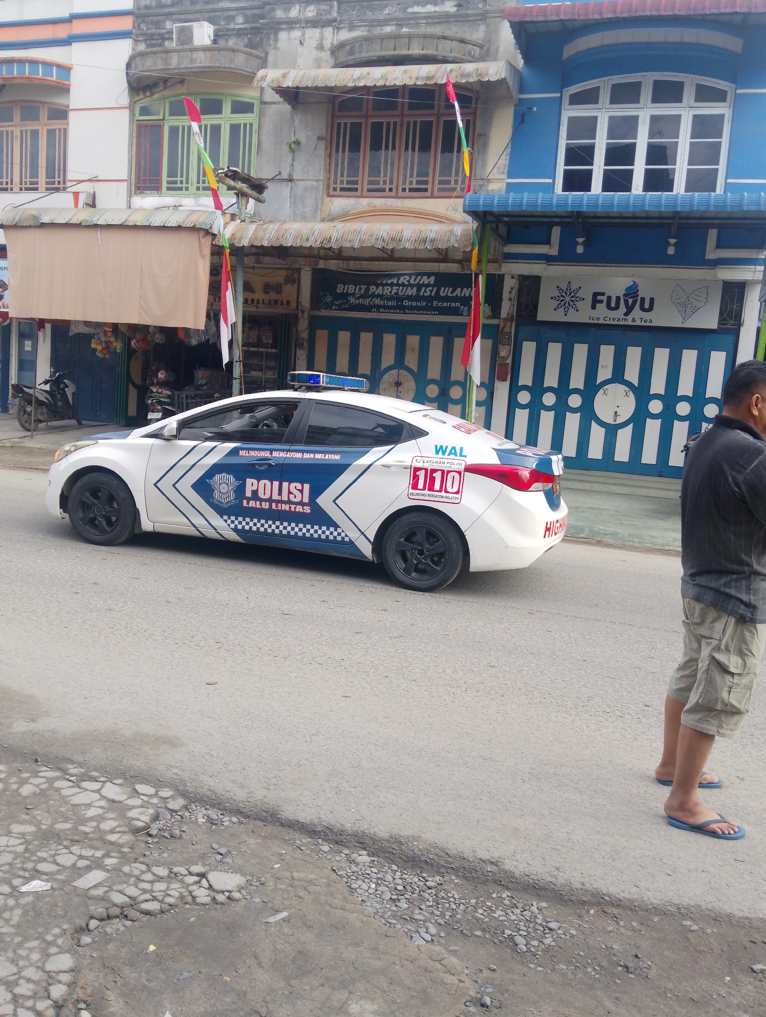 In the city of Serbelawan, Indonesia, a white police car with blue and white markings is parked on an asphalt road. The car has "POLISI LALU LINTAS" written on its side. A man wearing a dark t-shirt and light-colored shorts stands to the right of the car. In the background, there are buildings with colorful awnings and flags. One building has a sign that reads "Fuyu Ice Cream & Tea." The road appears to be in disrepair, with potholes and cracks visible.