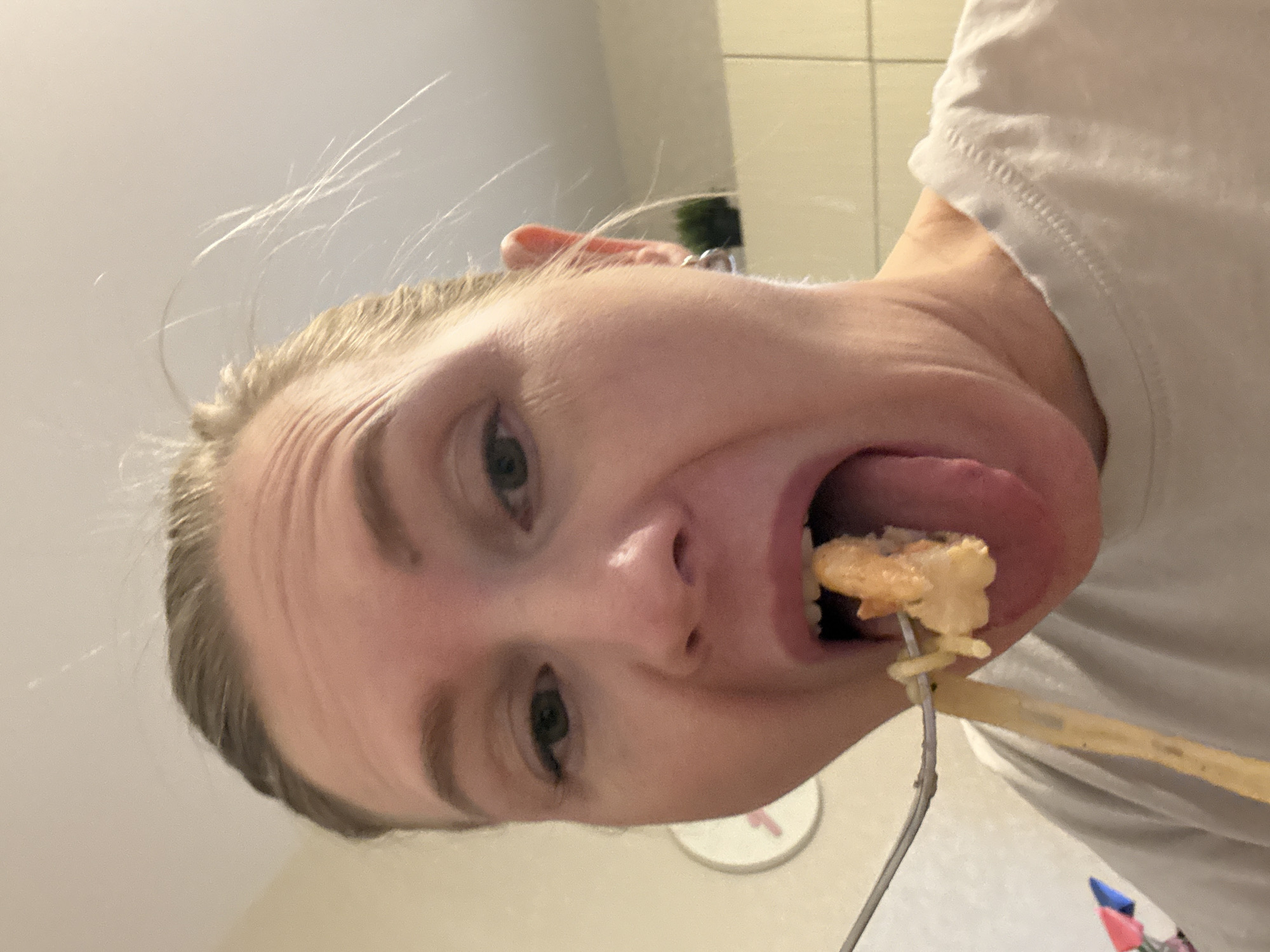 A close-up, distorted selfie of a fair-skinned person with blond hair and green eyes. The person is holding a fork with food impaled on it and is positioning it in their mouth. The person is wearing a white shirt. The background is a light-colored wall. The location is not identifiable as Yekaterinburg, Russia, from the given image.
