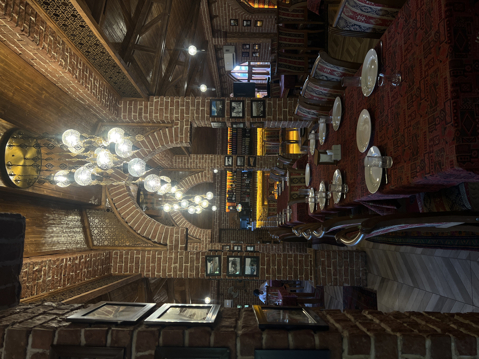 The image shows the interior of a restaurant in Şəki, Azerbaijan. The restaurant features exposed brick walls, wooden beamed ceilings, and arched doorways. The tables are set with red patterned tablecloths, white plates, and glassware. Several ornate chandeliers hang from the ceiling, illuminating the dining area. In the background, a bar area with shelves of bottles is visible, along with a computer monitor and an air conditioning unit. A single person is seated at a table in the distance. The overall ambiance is one of traditional architectural style combined with modern restaurant amenities.