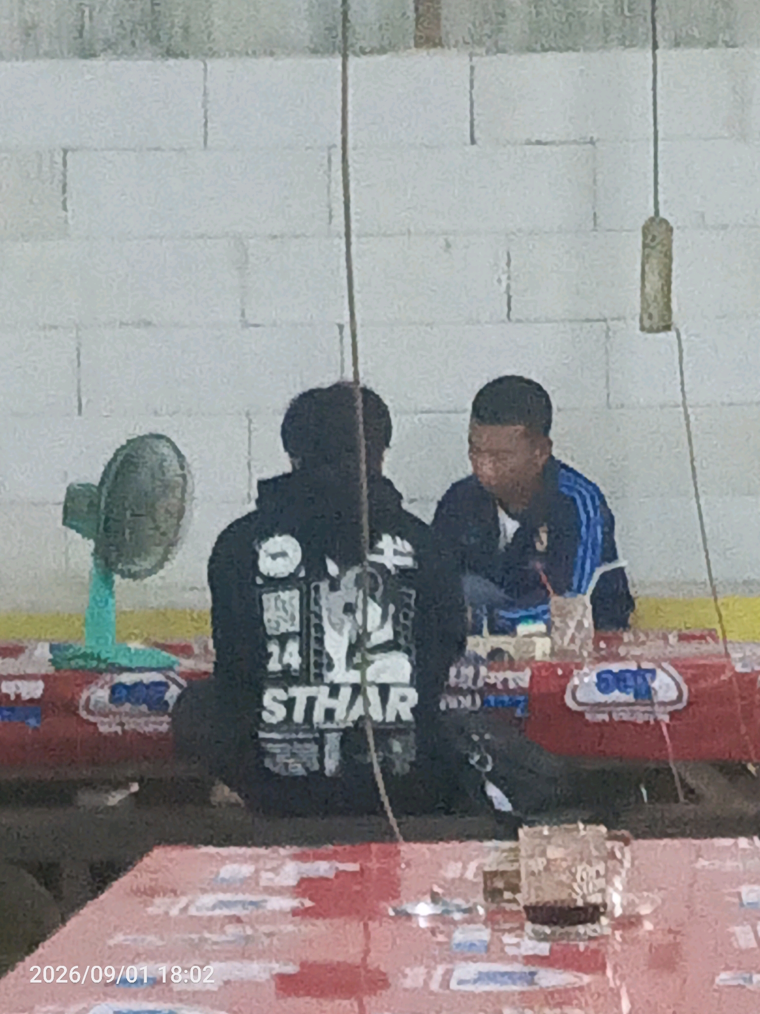The image captures an outdoor scene, likely in the city of Sidokerto, Indonesia, showing two individuals seated at a table. The foreground features a red table with a date and time stamp "2026/09/01 18:02" in the bottom left corner. Behind the table, a man on the right wears a blue and black track jacket with white stripes and appears to be speaking, with his mouth open. To his left, another person in a black hoodie with a graphic design on the back is seated. The hoodie's design includes the text "STHAR" and the number "24". To the left of the seated individuals is a turquoise desk fan. The background consists of a white brick wall and a brown hanging object. Red and white packaging or banners are visible on the table in front of the seated individuals.