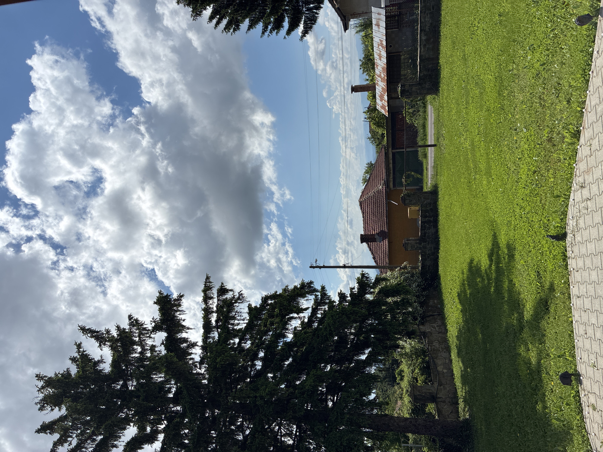 The image depicts a rural scene in Kormiansko, Bulgaria. A yellow building with a red-tiled roof and a chimney stands to the left, behind a low stone wall partially obscured by foliage. An adjacent building, partially visible, has a corrugated metal roof. A verdant grassy lawn stretches out, marked by the shadows of trees. A paved walkway borders the right edge of the frame. Overhead, power lines extend across a sky filled with a mix of blue and white, puffy clouds. Trees frame the upper and lower portions of the photograph.