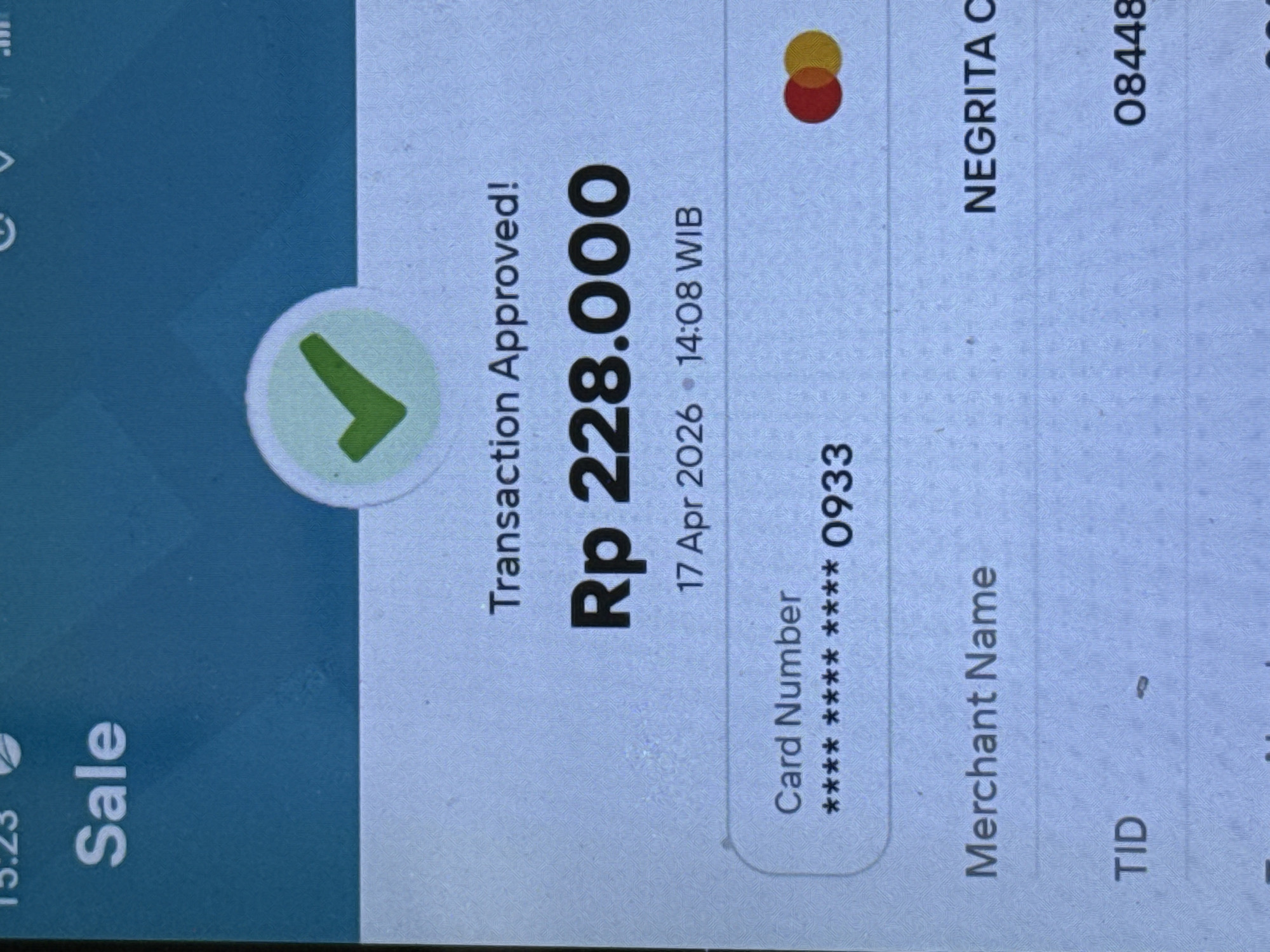 This close-up image captures a digital screen, likely a point-of-sale (POS) terminal or a smartphone, displaying a successful transaction in North Kuta, Indonesia. The screen shows a "Transaction Approved!" message with a prominent green checkmark icon. The main detail highlighted is the transaction amount: Rp 228.000 (Indonesian Rupiah).
Further details include the transaction date and time, "17 Apr 2026" at "14:08 WIB" (Western Indonesia Time). The card used for the purchase is partially masked, showing "Card Number **** **** **** 0933". A Mastercard-like logo is visible at the top right. The merchant's name is partially displayed as "NEGRITA C", and a "TID" (Terminal ID) of "08448" is also visible. On the far left, "Sale" is indicated, along with a device time of "15:23". The screen has a clean interface with a blue and light grey color scheme. The image is a direct photograph of a screen, showing slight glare and pixellation.