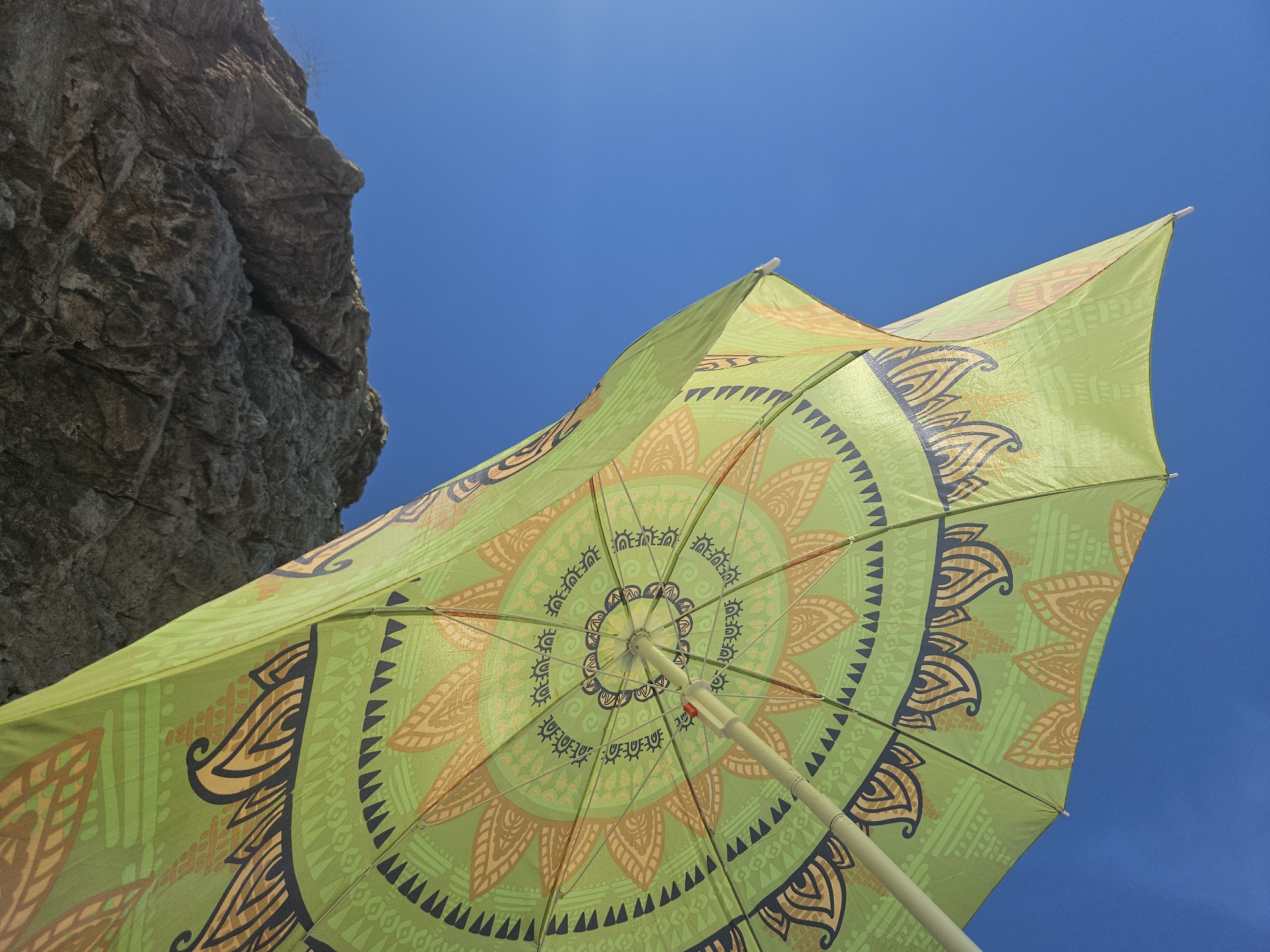 An underside view of a green, patterned beach umbrella is captured against a clear blue sky. The umbrella features intricate designs in orange and dark blue, including circular motifs and wave-like patterns. A portion of a large, textured rock formation juts into the frame from the upper left corner, casting a slight shadow on the umbrella. The scene appears to be taken from a low angle, looking upwards. The image likely depicts a sunny day at a beach in Čanj, Montenegro. There are no people visible in the frame.