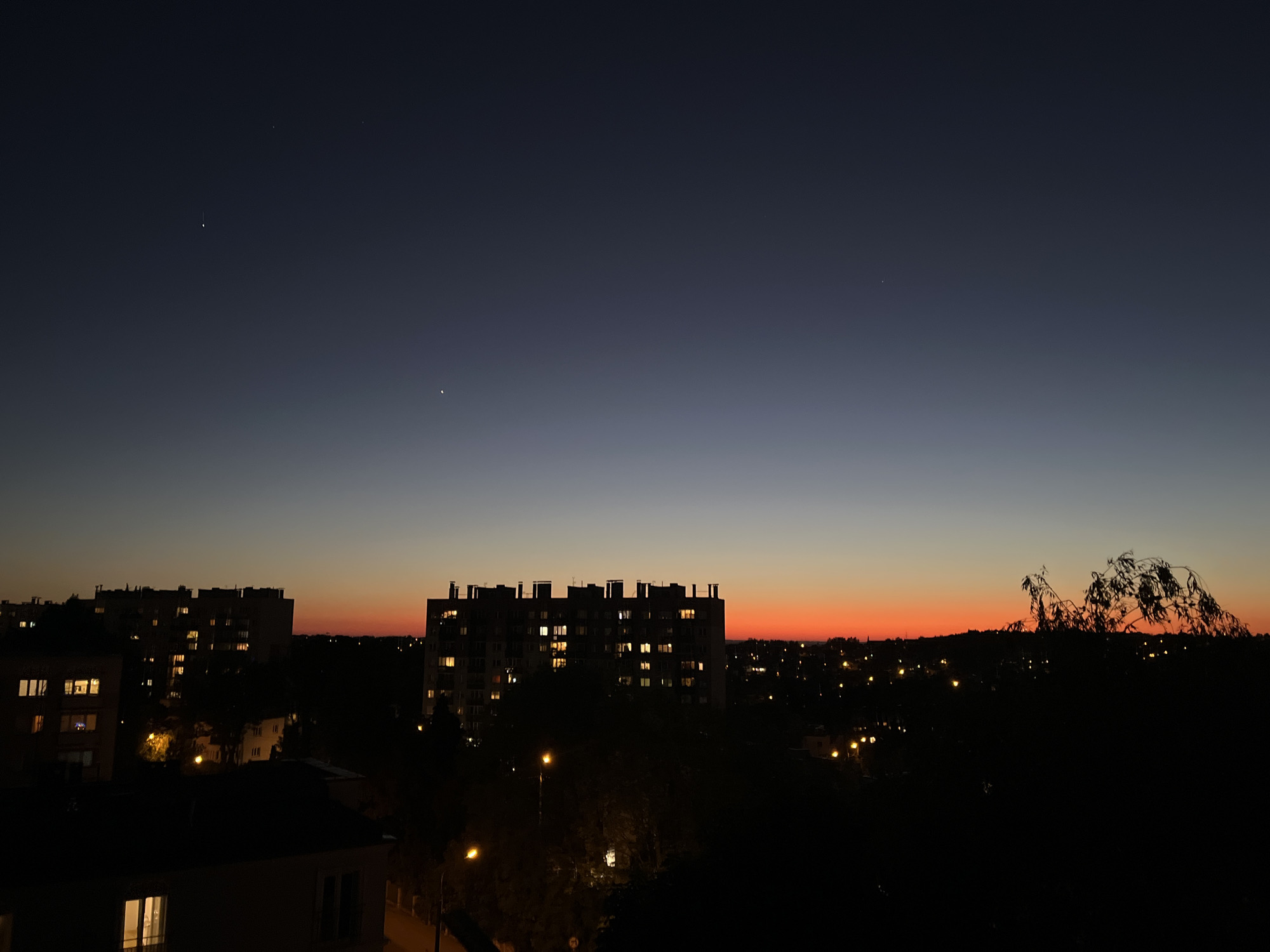 The image shows a cityscape at dusk or dawn. In the foreground, a few buildings are visible with some illuminated windows, indicating it is either very early morning or late evening. Several streetlights are lit, adding to the illumination. The central part of the image features a tall, multi-story building with numerous lit windows, suggesting residential occupancy. Beyond these buildings, a visible gradient in the sky ranges from dark blue at the top to orange and red near the horizon, indicative of a sunrise or sunset. The horizon is partially obscured by trees and other buildings. There are a few faint stars or planets visible in the sky. The location is inferred to be urban, but specific details are missing to definitively place it in a city of Unknown, Unknown. The image lacks any identifiable people or explicit actions.