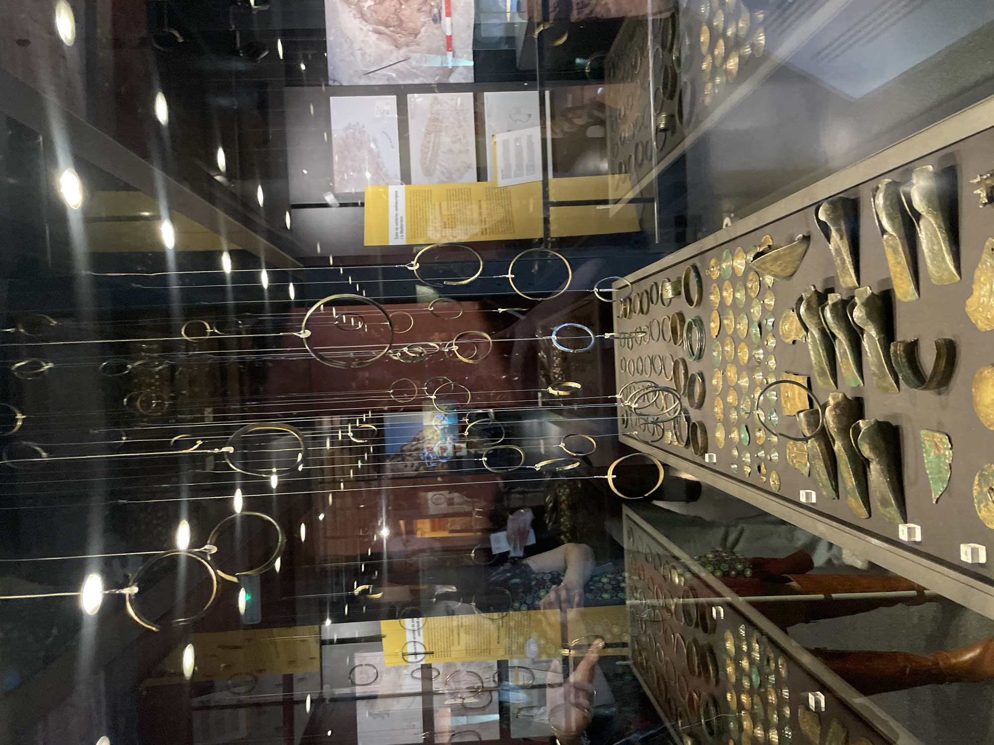 The image shows the interior of a museum, likely in Barcelona, Spain, given the cultural artifacts. The scene is viewed through a glass display case containing an array of ancient bronze tools and jewelry items, including axes and bracelets. Hanging in front of the display, suspended on wires, are many circular objects. These are likely representations of jewelry. The background features museum signage and other exhibits. The reflection in the glass case reveals a person looking at the artifacts.