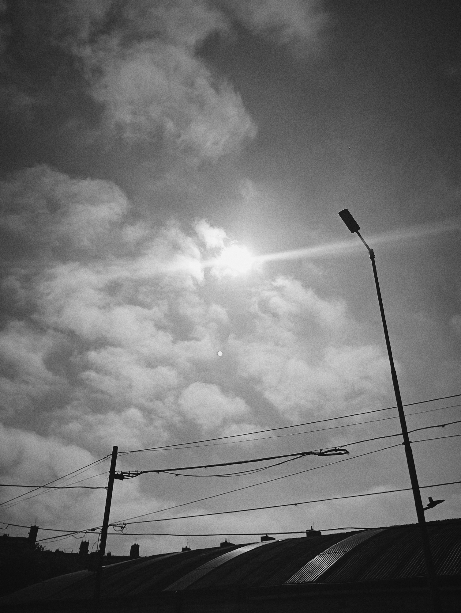 A black and white photograph depicts a sky filled with clouds and the sun. In the foreground is a street lamp, power lines and the corrugated metal roof of a building. There are also chimneys visible in the silhouette of the roof. The photograph's perspective is from below, looking upwards, capturing an urban landscape. The location cannot be determined without further information.