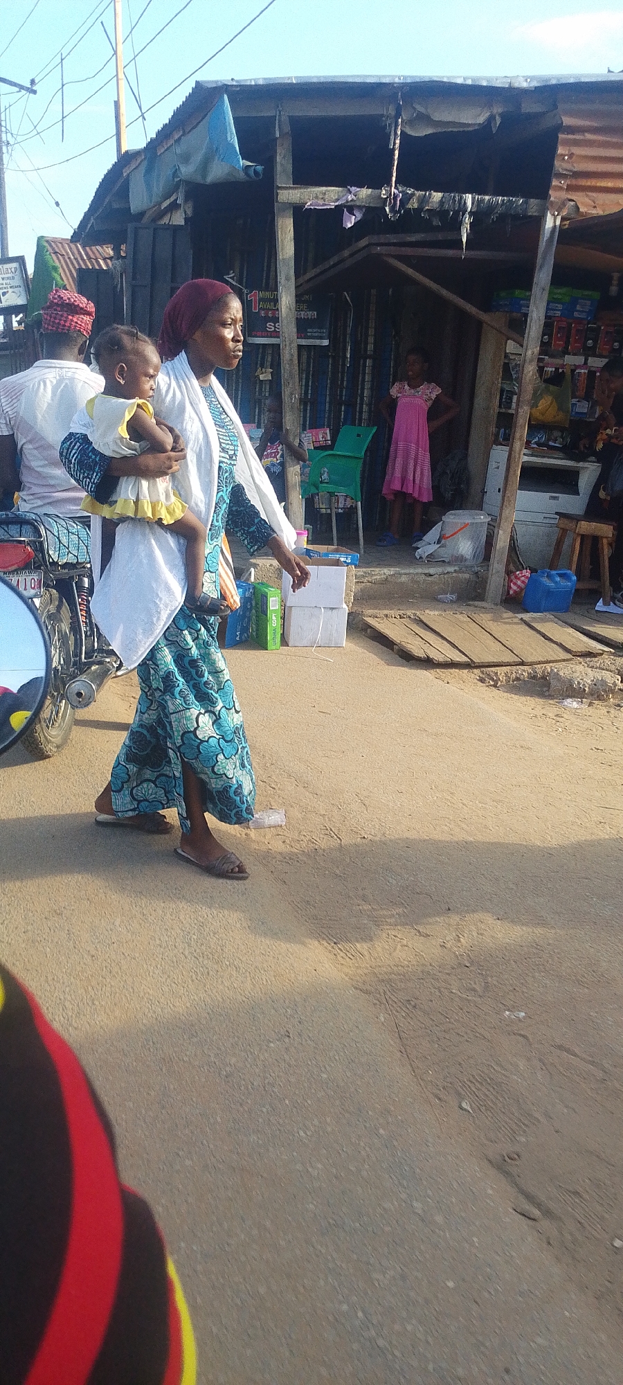 The image shows a woman carrying a child on a street in Lokoja, Nigeria. The woman is wearing a blue and teal patterned dress with a white shawl and a maroon headscarf. She is walking on a paved road near a building that appears to be a small shop or store. A girl in a pink dress is standing inside the shop. There is a man with a red and white patterned head covering on a motorcycle next to the woman. The shop has various items for sale, including electronics and drinks. The environment seems urban with basic infrastructure.