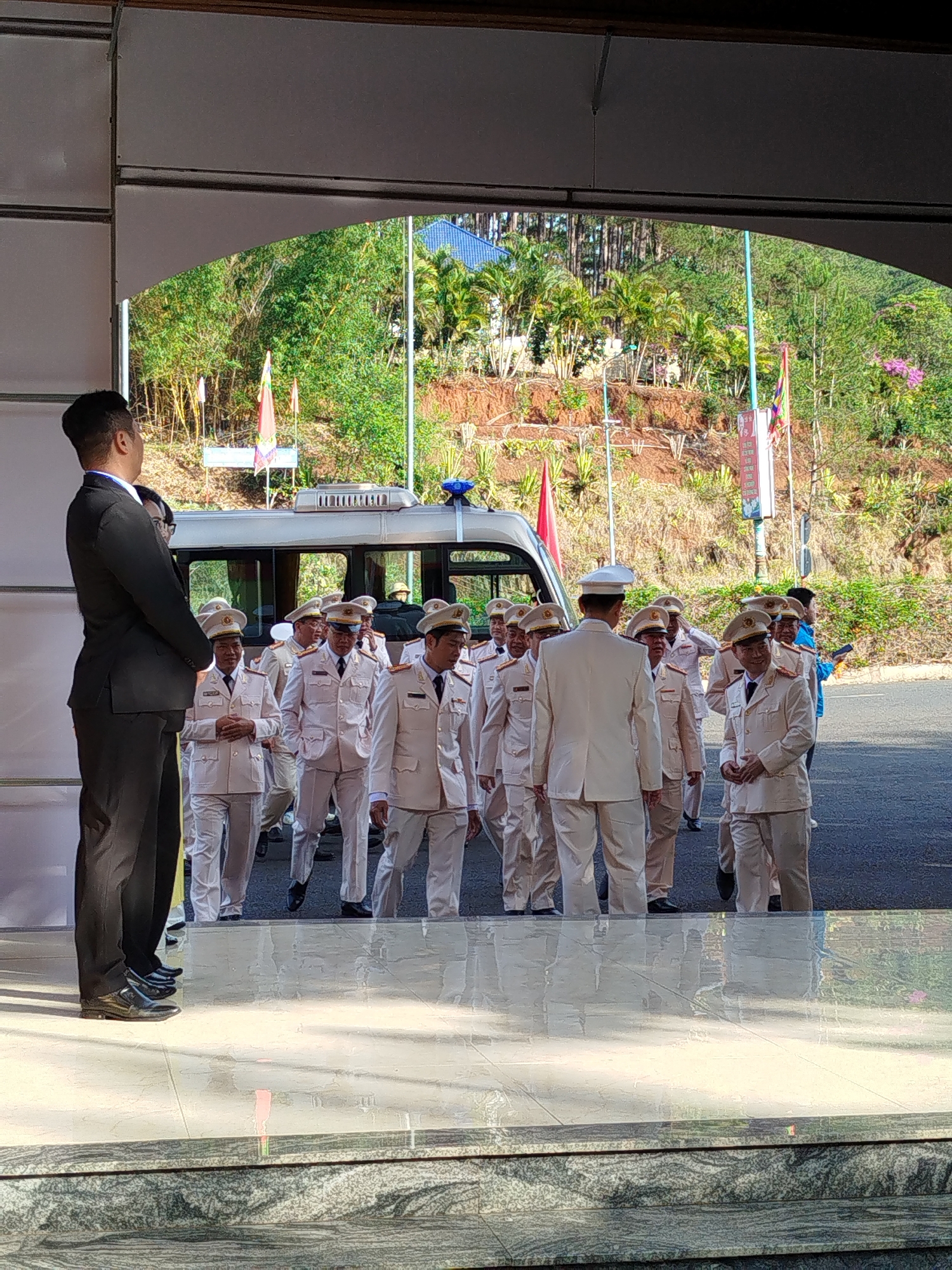 A group of approximately 14 people in light tan uniforms with hats are walking towards the viewer through a building opening. A black van with a blue light bar is visible behind them. A man in a dark suit stands with his back to the viewer to the left of the opening. The background includes greenery, flags, and buildings. The scene appears to be in Da Lat, Vietnam, based on the landscape and style of buildings and flags.