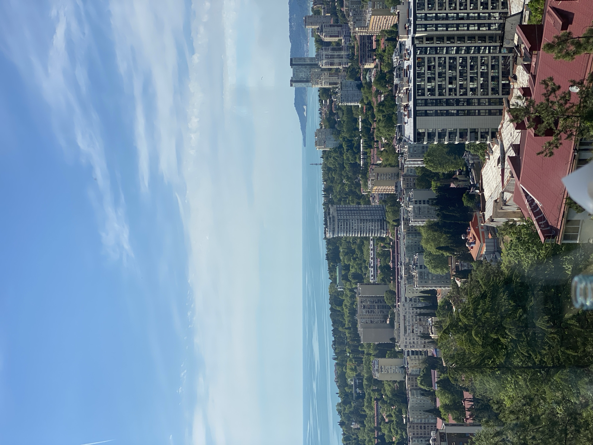 This image captures a panoramic view of the coastal city of Sochi, Russia, looking out over the Black Sea under a bright, clear sky. The scene is oriented correctly as a landscape, though the provided image is rotated.
The top portion of the image features a vast blue sky, dotted with sparse, wispy white clouds, indicative of fair weather. A faint airplane contrail is visible in the lower left area of the sky. Below the sky, a wide expanse of calm, light blue water stretches across the middle of the frame, representing the Black Sea. The distant horizon line is clearly visible.
Along the right side of the frame, bordering the sea, lies the city's densely built coastline. This urban landscape is characterized by a mix of buildings: numerous multi-story structures, including modern high-rises with many windows and older buildings, some featuring distinctive red-tiled roofs. These structures are interspersed with abundant lush green trees and foliage, creating a vibrant contrast between urban development and natural greenery. In the far distance, faint outlines of mountains or hills can be seen merging with the hazy sky above the sea, adding depth to the panorama.
In the immediate foreground on the right, parts of a red-tiled roof and green tree branches are visible, suggesting the photo was taken from an elevated vantage point like a balcony or terrace. A blurred, transparent, round object, possibly a glass, is partially seen on the far right. The overall lighting suggests it is daytime, likely morning or early afternoon, with high visibility and no discernible human activity other than the distant presence of the city itself. No text is visible in the image.