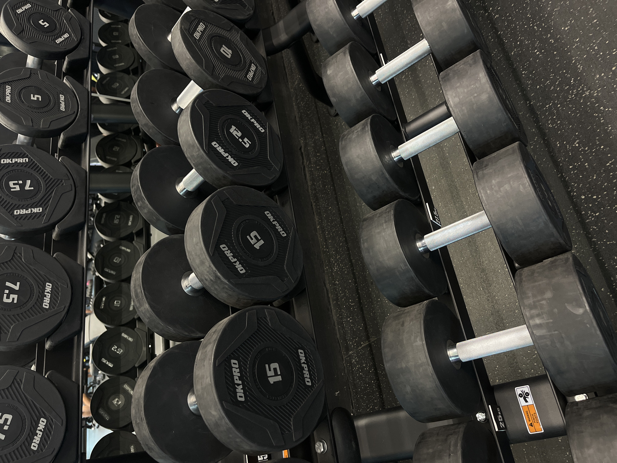 This image displays a gym interior, focusing on a multi-tiered rack filled with various dumbbells. The scene is brightly lit, showcasing rows of black dumbbells with circular weight plates and shiny silver knurled handles. The dumbbells are arranged on sturdy black metal racks, suggesting an organized fitness environment. The floor beneath the rack is dark, possibly a rubberized gym flooring, with subtle white speckles.
The dumbbells prominently feature the brand name "OKPRO" and their weight in white numerals. Visible weights include 5, 7.5, 10, 12.5, and 15 units. A warning sticker is affixed to one of the rack supports, displaying "▲ WARNING", "PINCH POINTS", a pictogram of crushing hands, and the text "25KG". Reflections in the background show additional dumbbells, including one with "2.5" visible. The dumbbells are neatly racked, indicating the area is set up for weightlifting and strength training activities.
Location context: Unknown, Unknown