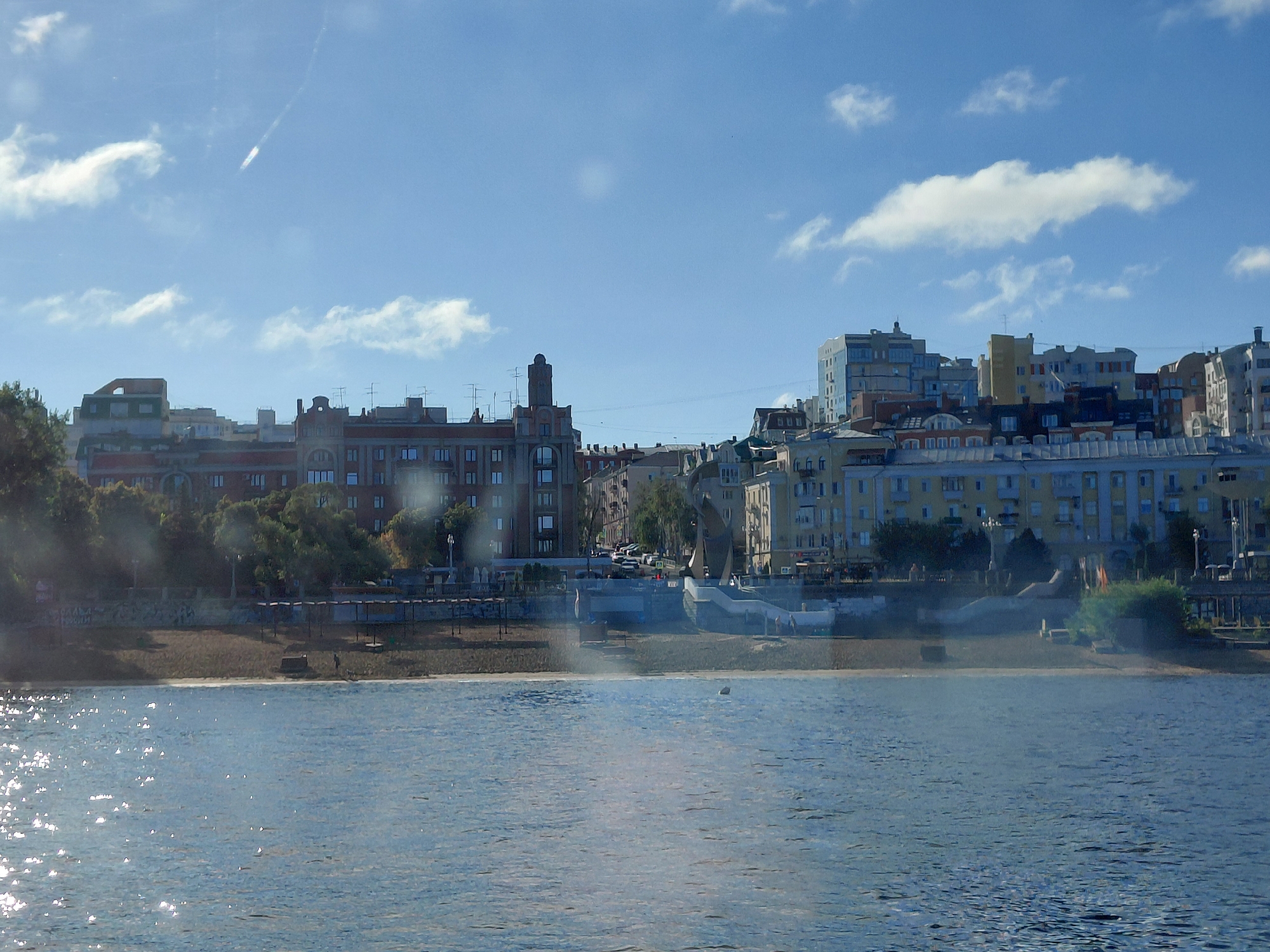The image depicts a wide river in the foreground, reflecting the sunlight, which creates shimmering patterns on the water's surface. Along the riverbank, a sandy shore transitions into a tiered embankment adorned with trees and urban structures. Behind the embankment, a cityscape rises, characterized by a mix of architectural styles. Buildings vary in height and color, with some featuring red brick facades, while others are painted in lighter hues like yellow and blue. Rooftops display antennas and some have visible window panes. Infrastructure such as streetlights and power lines are discernible. A pathway or promenade runs along the riverbank, bordered by trees and shaded areas. Several vehicles are parked along the road behind the embankment. The sky is a clear blue, interspersed with scattered white clouds, one of which is long and streaky. A faint streak of light, possibly from an aircraft, is visible in the upper left portion of the sky. The scene suggests a daytime view of a waterfront urban area in Verhnyaya Podstyopnovka, Russia, likely during a sunny period.