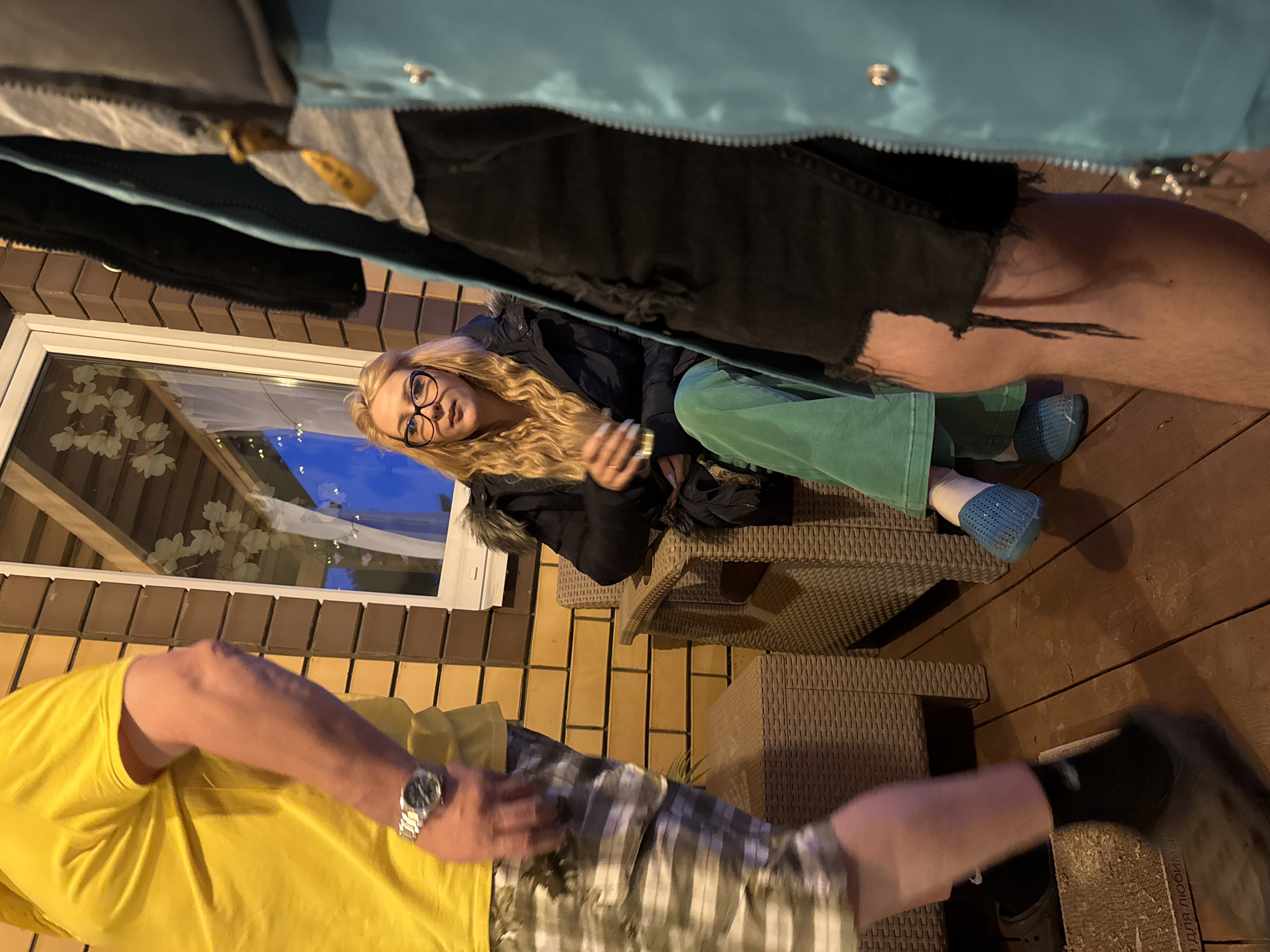 The image depicts an outdoor gathering at night, likely on a porch or deck, with brick and wood structures visible. In the foreground, a person in a yellow t-shirt and patterned shorts is partially visible on the left. To the right, a person wearing a light blue jacket and black ripped jeans is also partially visible. Seated in a wicker chair in the middle ground is a blonde woman with glasses, wearing a dark puffer jacket and green pants, holding a beverage. The background shows a window with some decorations, and the overall lighting suggests it is evening or night. The location is identified as Большая Венья, Russia.