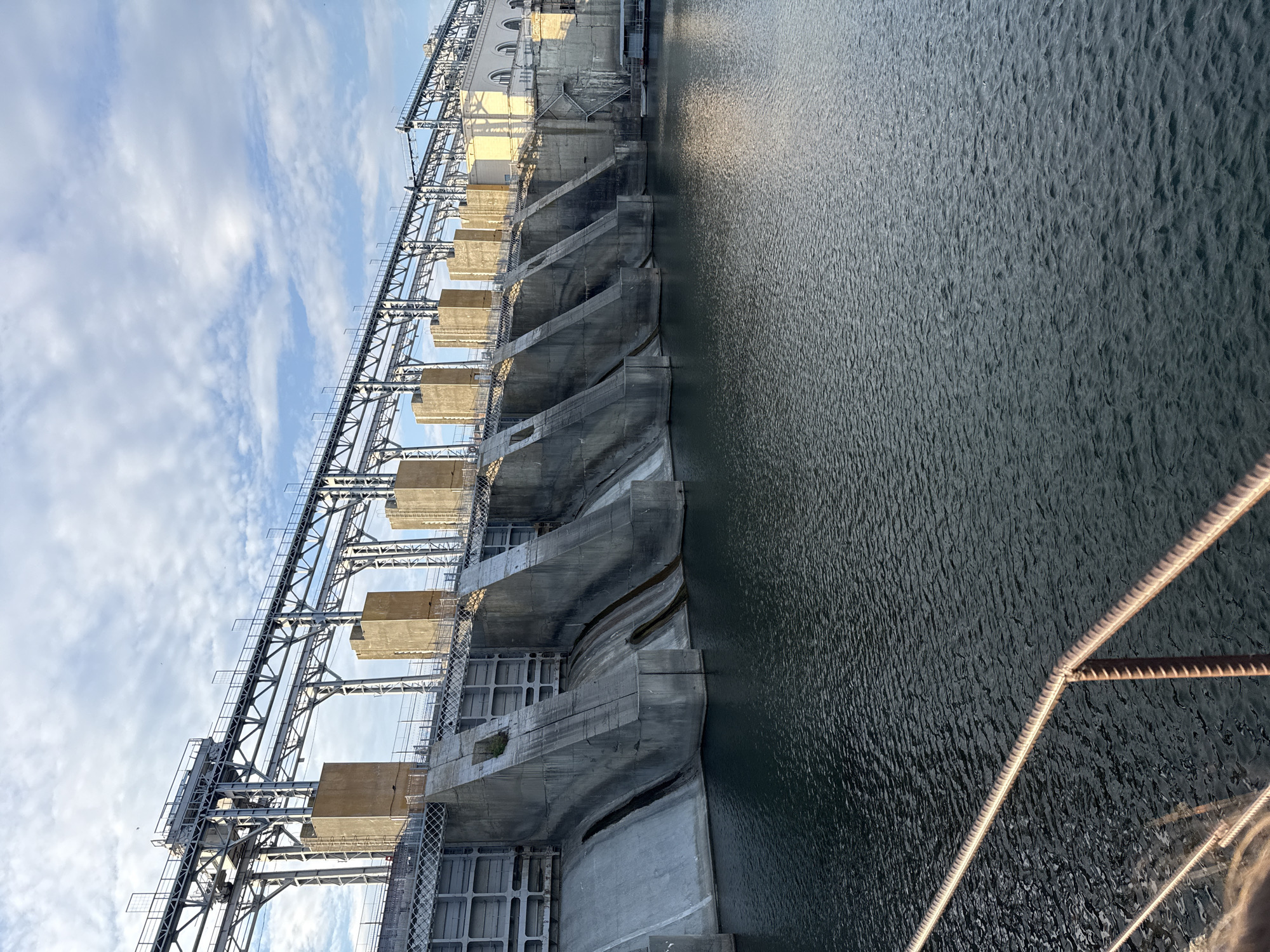 A large concrete dam with spillways is visible, with a metal superstructure above. The water in front of the dam is dark and rippled. In the foreground, a metal railing runs horizontally across the image. The sky above is partly cloudy. The scene is static, with no visible people or actions. The dam is located in the city of Ustia, Moldova.