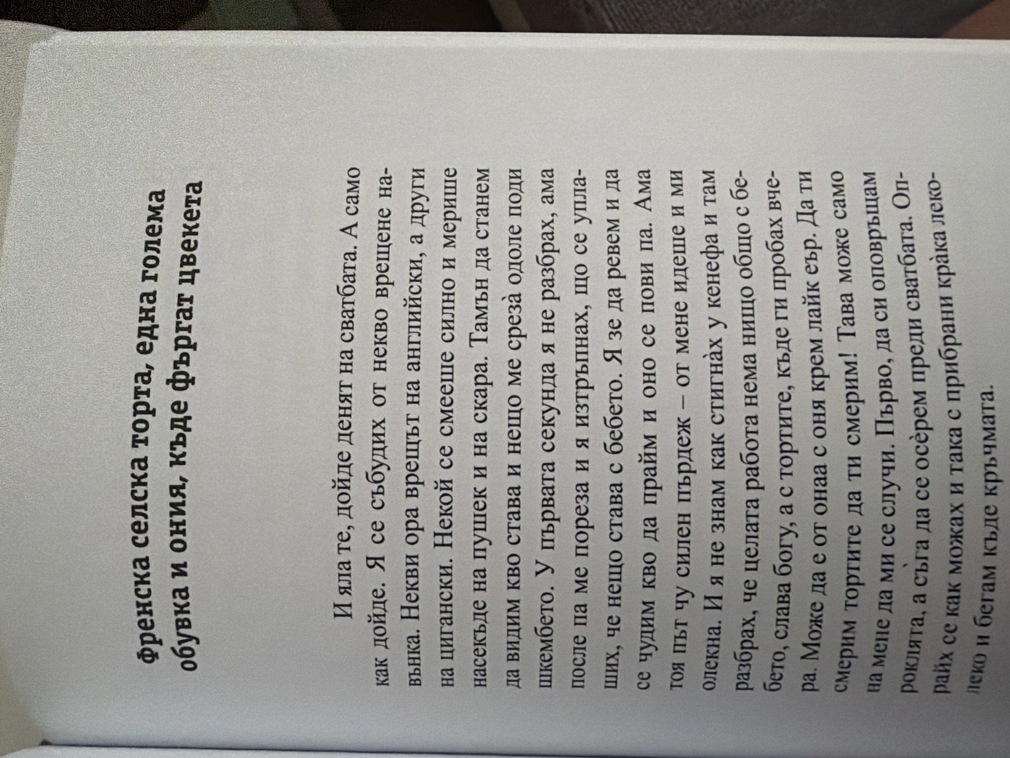 The image contains a page from a book with Bulgarian text. The text discusses a "French village cake" and a story about waking up to the sounds of someone shouting in English and other languages. The story then shifts to a situation involving a baby and a strong smell, leading to the narrator to run out of the house. The scene appears to be indoors, likely in a room where the book is being read. There is no indication of any specific location in Pazardzhik, Bulgaria.