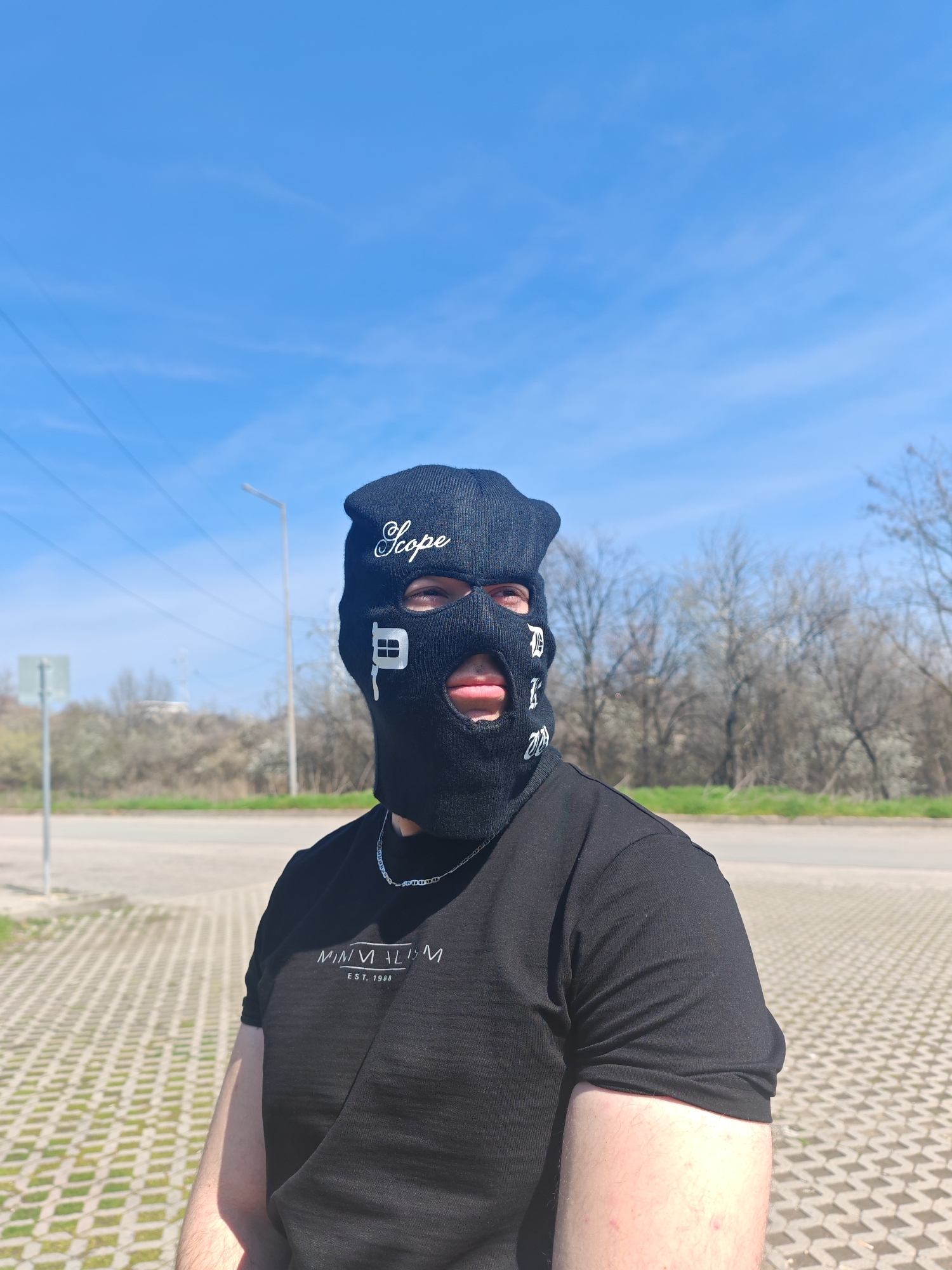 A male individual is depicted outdoors in what appears to be an urban-edge or industrial-zone area within Shumen, Bulgaria. The exact location is not discernible from the image. The subject, positioned slightly right of center, faces slightly left while looking directly towards the camera. He wears a black balaclava covering his head and face, emblazoned with white text: "Scope" in a cursive font across the forehead, and multiple stylized "P" characters vertically on the right side (viewer's perspective) of the face opening. His eyes are visible through the eyeholes, and his mouth and chin are exposed through the mouth opening, revealing short facial hair on his chin. He wears a black short-sleeved t-shirt with white text on the front reading "MINIMALISM EST. 1988". A silver-colored chain with anchor links is visible around his neck. His left arm is visible, showing pale skin.
The foreground consists of a light-colored paved surface made of rectangular concrete pavers arranged in a grid pattern, with sparse green grass visible between some pavers. Behind the subject, a darker paved road or parking area is present, bordered by a strip of green grass. Beyond the grass, a line of leafless deciduous trees and shrubs is visible, consistent with a late winter or early spring season. To the viewer's left, a tall, light-colored streetlamp post stands prominently, with multiple overhead power lines extending horizontally across the mid-ground. A smaller, indistinct gray signpost is also present further left. In the far background, a gentle hill covered in bare trees and an indistinct light-colored building structure are visible. The sky is clear and bright blue with minimal wispy clouds, indicating a sunny daytime environment.