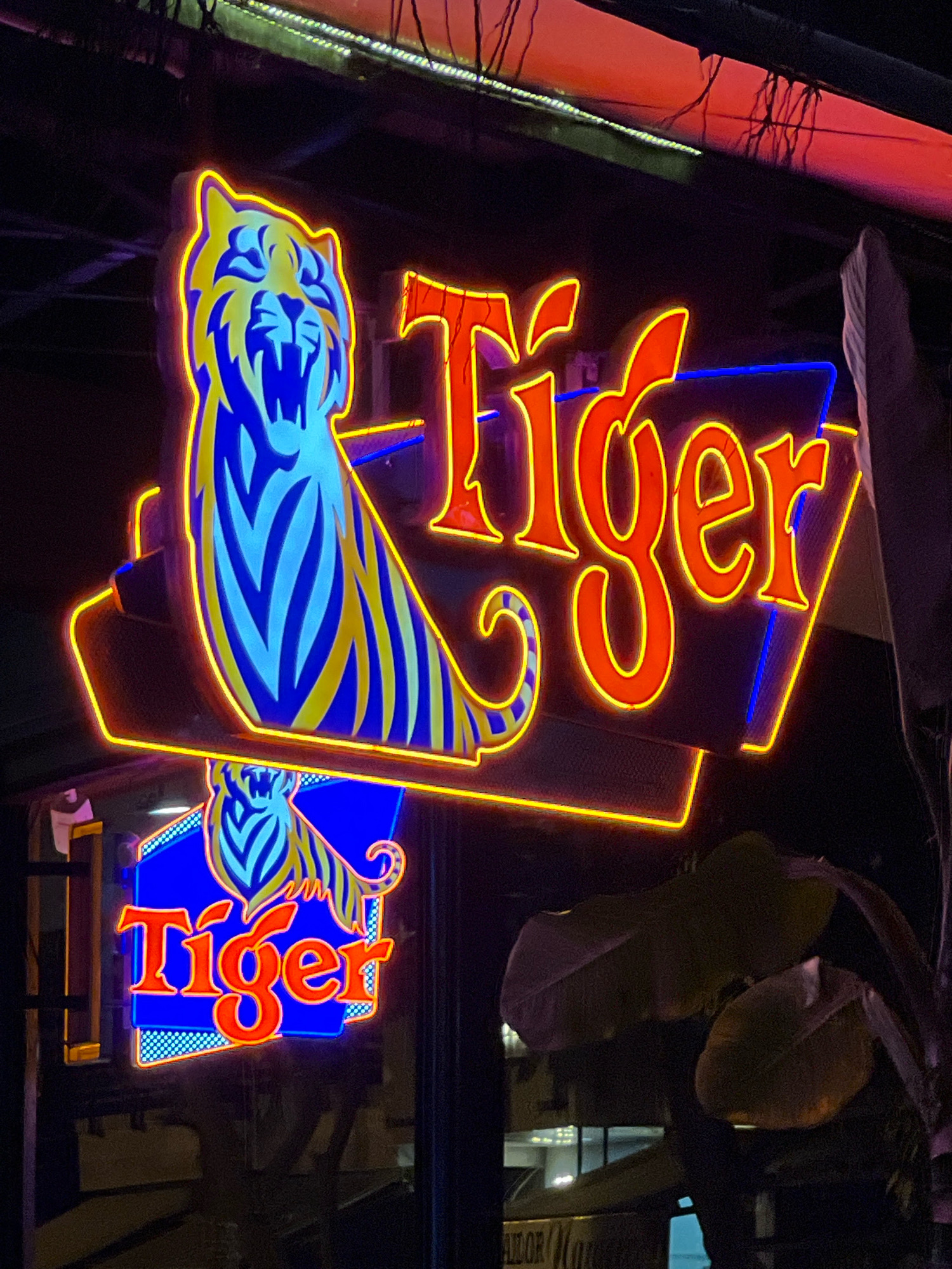 The image shows a neon sign for "Tiger" beer, featuring a stylized tiger's head and body in blue and yellow. The sign is illuminated with orange and yellow neon outlines. Below this larger sign, a smaller, identical "Tiger" sign is visible through a window, also with the tiger graphic and the word "Tiger" in orange neon. This smaller sign appears to be on a glass surface, reflecting the surroundings. The overall setting is dark, suggesting nighttime, with hints of other lights and foliage visible in the background. The neon lights are vibrant against the dark backdrop. This specific Tiger beer neon sign is located at a establishment on Tran Phu Street in Nha Trang, Vietnam.