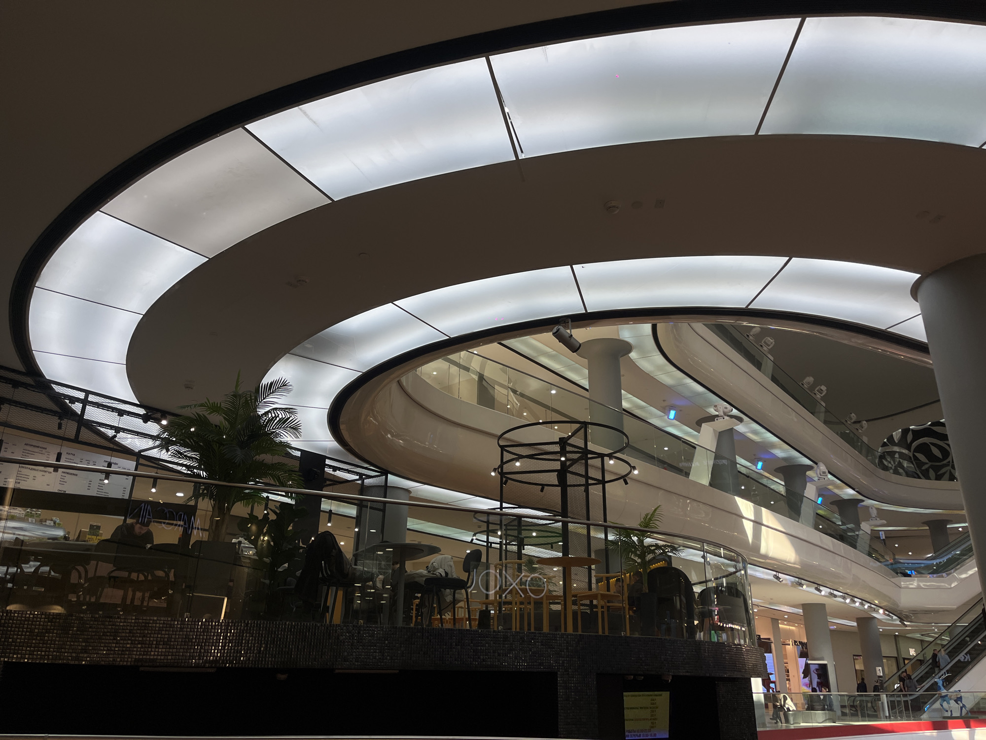 This media file captures the interior of a modern, multi-level shopping mall or commercial complex in Krasnodar, Russia. The perspective is from a lower floor, looking up and across the expansive space.
The dominant architectural feature is a series of sweeping, curved balconies and walkways, defined by glass railings and smooth, light-colored surfaces. Above, a large, curved ceiling is composed of illuminated rectangular panels, providing bright, diffused artificial light throughout the structure. Escalators are visible on the right side of the frame, connecting the various levels, with a few people discernible on them.
In the mid-ground, a cafe or restaurant area is situated on an elevated platform. This space features several tables and chairs, a counter, and a distinctive black, open-frame structure adorned with small lights. A large palm plant and other greenery add a natural element to the setting. A few individuals are seated at tables in the cafe. Behind the counter, a menu board with Cyrillic text is visible, and the word "JOKO" is displayed on the back wall of the establishment.
Further down on a lower level, another sign with Cyrillic text is faintly visible. The overall scene depicts a bustling, yet orderly, commercial environment, characteristic of a large indoor shopping center, lit primarily by artificial sources.