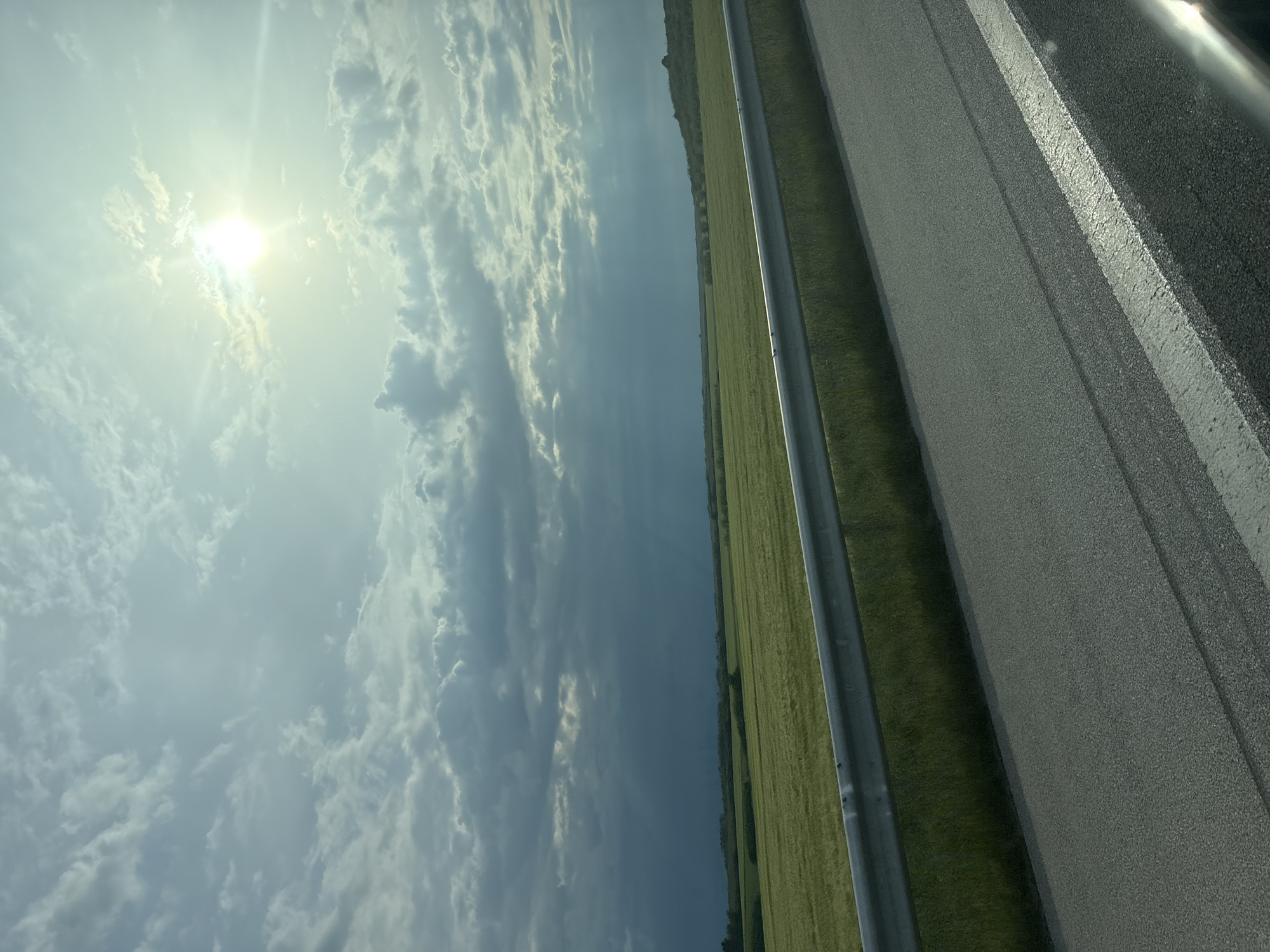 The image depicts a view of a sky with scattered clouds and a bright sun, alongside a stretch of green field, a metal guardrail, and a paved road. The road has a white painted line. The image appears to be taken from a moving vehicle. There is no evidence to definitively place the location in Zetiovo, Bulgaria.