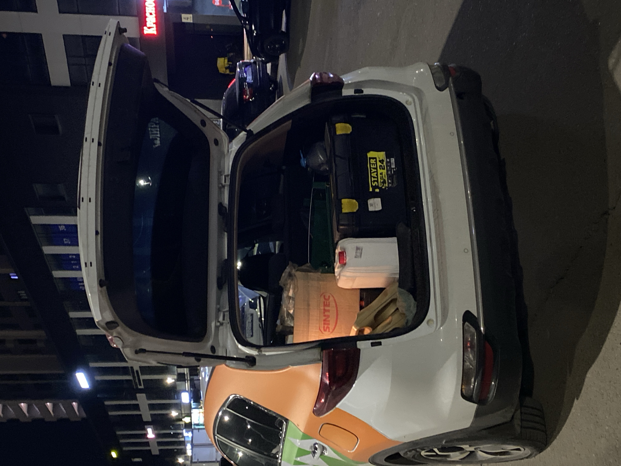 A white car, possibly a hatchback or small SUV, with an orange and green livery on its side, is parked on a paved surface at night with its trunk wide open. The image is rotated, showing the car on its side.
Inside the open trunk, several items are visible. A black tool case with yellow latches bears the text "STAYER" and "SA 24". Next to it is a white plastic jerrycan or container with a red cap. Further within the trunk, a cardboard box is visible with the brand "SINTEC" printed in red. Other unidentified items, including what appears to be fabric or bags, are also stowed.
The background features illuminated buildings, indicating an urban or commercial setting at night. A prominent red illuminated sign on one building partially displays "КРАСНОО" (likely part of a Russian word like "КРАСНОЕ"). Reflections of other buildings and lights are visible in the car's surfaces. Another vehicle is partially seen in the background. The scene is lit by artificial light sources from the surrounding environment.