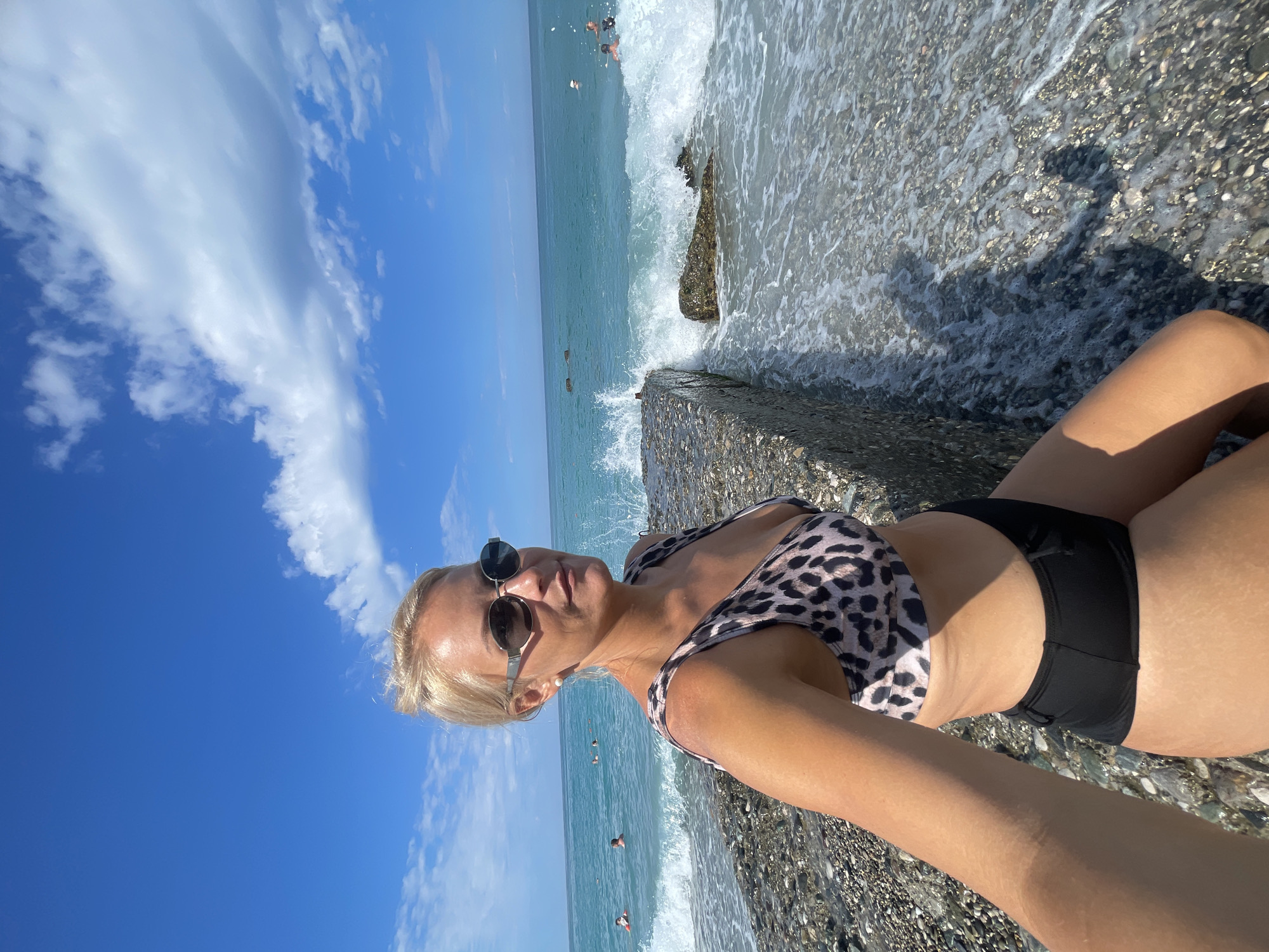 The image shows a blonde woman in a leopard-print bikini and black bottoms, posing for a selfie on a rocky beach. She is wearing sunglasses and a pearl earring. The ocean is behind her, with waves crashing on a concrete pier. Several people are visible swimming in the turquoise water. The sky is blue with scattered white clouds. The location is identified as Gagra, Abkhazia.
