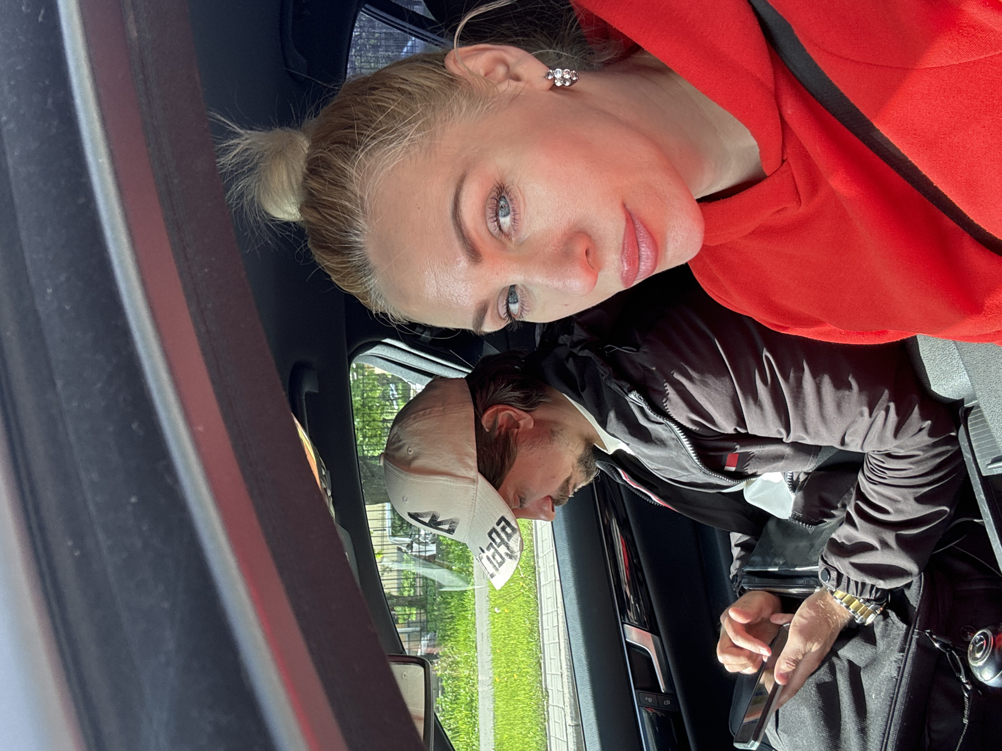 Inside a car, a blonde woman in a red hoodie looks towards the camera. Seated beside her, a man in a cap is looking down at a smartphone. Outside the car window, vegetation and a sidewalk are visible. The location is unknown.