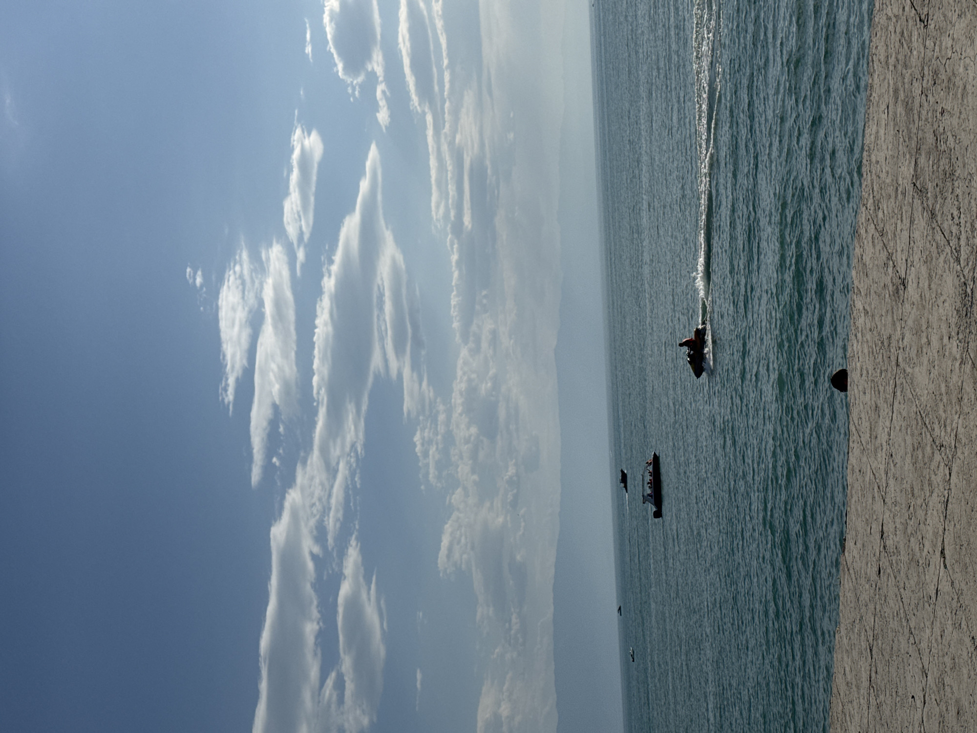 The image depicts a tranquil scene on the Caspian Sea near Zharbulak, Kazakhstan. In the foreground, a stone embankment runs along the bottom of the frame. The sea is a deep turquoise, with gentle ripples across its surface. Several boats are visible on the water. One jet ski is in motion, leaving a white wake behind it, and a person is riding it. Further out, there are other boats, including a larger vessel carrying passengers. In the distance, a hazy mountain range forms the horizon line, with a clear blue sky overhead dotted with scattered white clouds. The overall impression is one of a peaceful day by the sea.