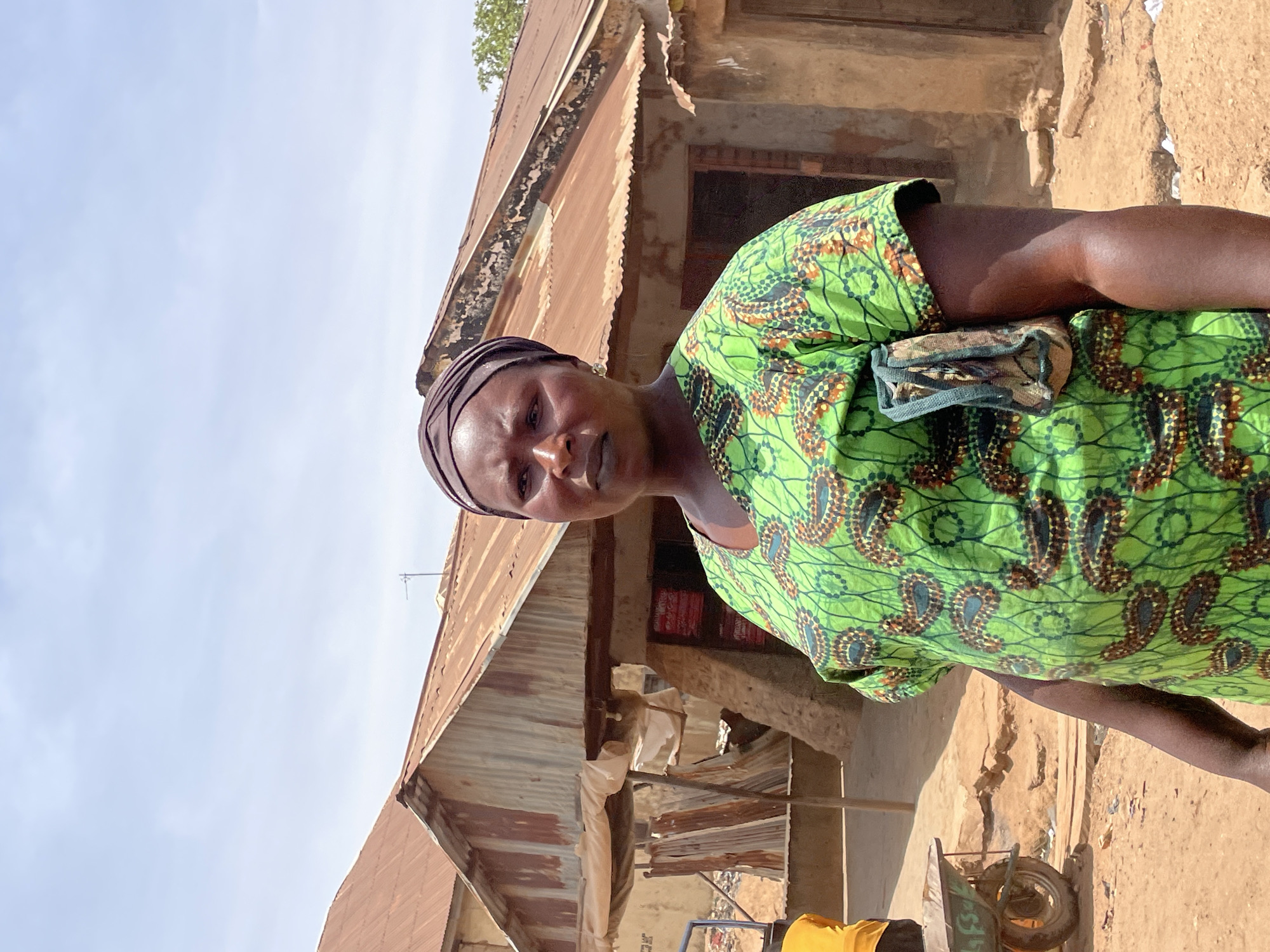 The image presents a direct portrait of a woman in an outdoor setting in Bukuru, Nigeria. She stands prominently in the foreground, looking directly at the camera with a neutral and composed expression. She has dark skin and is attired in a vibrant, short-sleeved green top featuring an intricate pattern of brown and black paisley-like designs. A small, patterned fabric pouch, coordinating with her top, hangs from her left shoulder. Her head is covered by a dark brown or black headwrap.
In the background, a cluster of rustic buildings with corrugated metal roofs is visible. These roofs appear weathered and rusted in parts, while the building walls are a light brown, possibly constructed from mud or unpainted concrete. The sky above is clear and light blue, indicating a bright, sunny day, likely mid-morning or afternoon, with strong sunlight casting distinct shadows.
The ground around the structures is dry, dusty earth. In the lower right corner, a portion of a small, green wheelbarrow or cart is visible, showing two wheels and some indistinct green writing on its side. A patch of yellow fabric or garment is also partially visible in this area. The overall scene depicts a modest, possibly rural or semi-urban environment in a warm climate. The woman is simply posing for the photograph; no other specific activity is evident. The visible text on the cart is not clearly legible.