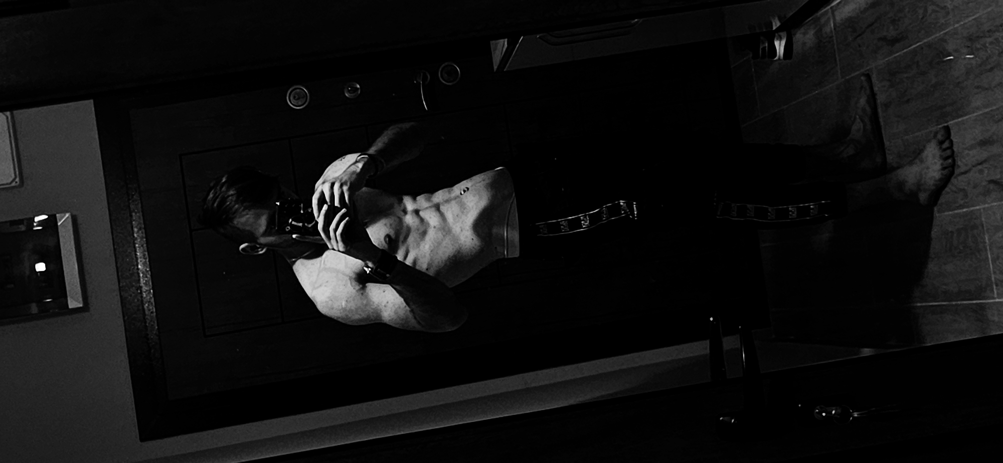 A black and white, high-angle shot depicts a shirtless man in a bathroom-like setting, likely in Plovdiv, Bulgaria. The man is positioned along a dark countertop or vanity, his head near one end and his bare feet extending towards the opposite end. He is holding something to his face with both hands. The surroundings include tile flooring, a mirror on a nearby wall, and various bathroom fixtures, such as sinks and taps, suggesting a domestic environment. The lighting is dramatic, with strong contrasts between light and shadow, obscuring details and emphasizing the subject's form.