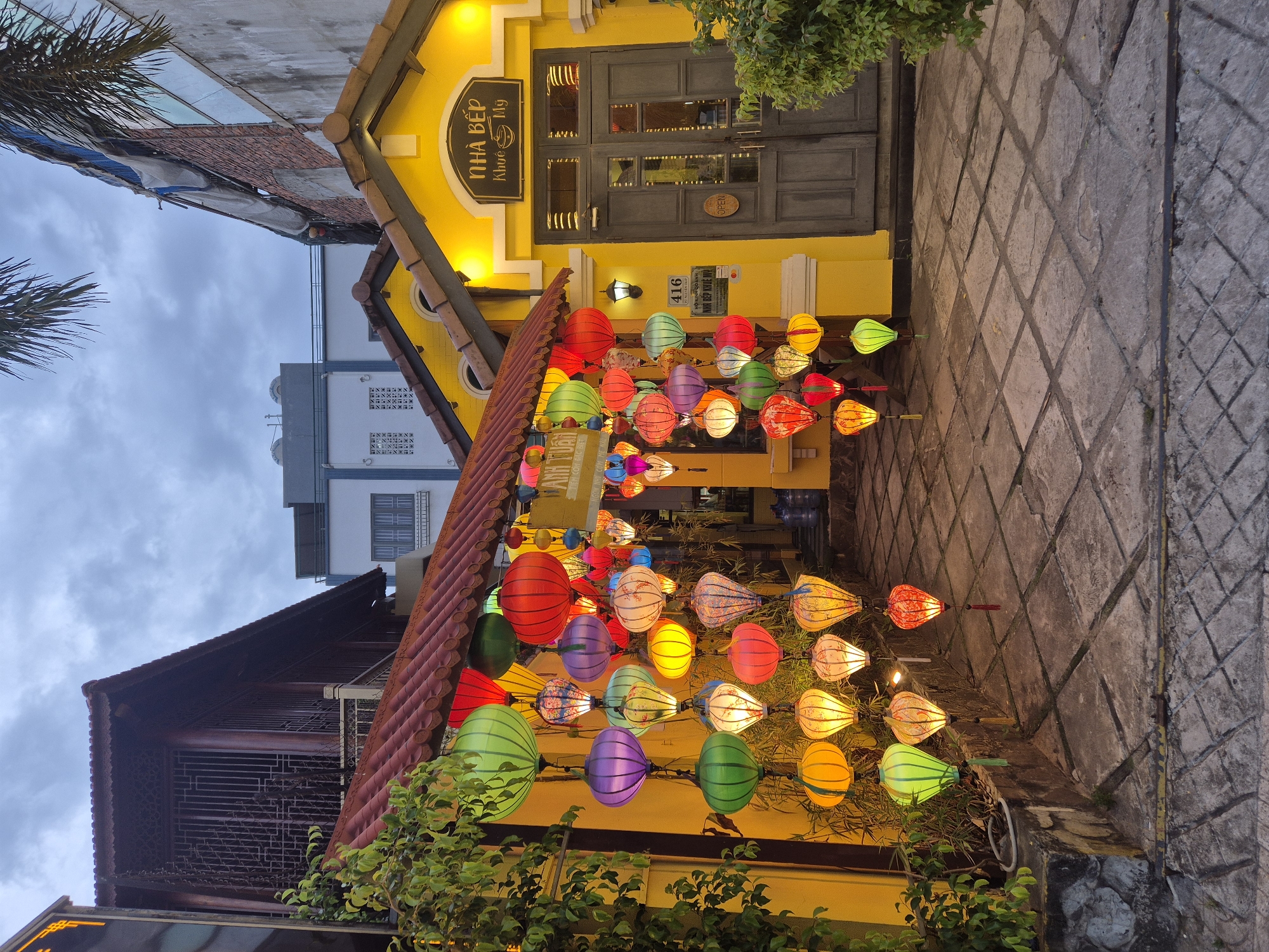 This is an exterior, eye-level, wide shot of a street in Da Nang, Vietnam, at dusk, featuring a bright yellow building adorned with numerous colorful, illuminated lanterns. The building has a traditional Vietnamese architectural style, with a tiled roof and wooden doors. A sign above the entrance reads "NHÀ BẾP Khuê Mỹ." The street is paved with large, square stone tiles. Green foliage and trees are visible on the left and right sides of the building. The sky is overcast and a pale gray, suggesting the beginning of evening. The lanterns, in various shapes and sizes, are strung in vertical arrangements and cascade down from the roofline of the building, creating a vibrant and festive atmosphere. There are no people visible in the scene. The lighting is primarily from the lanterns and a single wall-mounted light fixture next to a sign with the number "416."