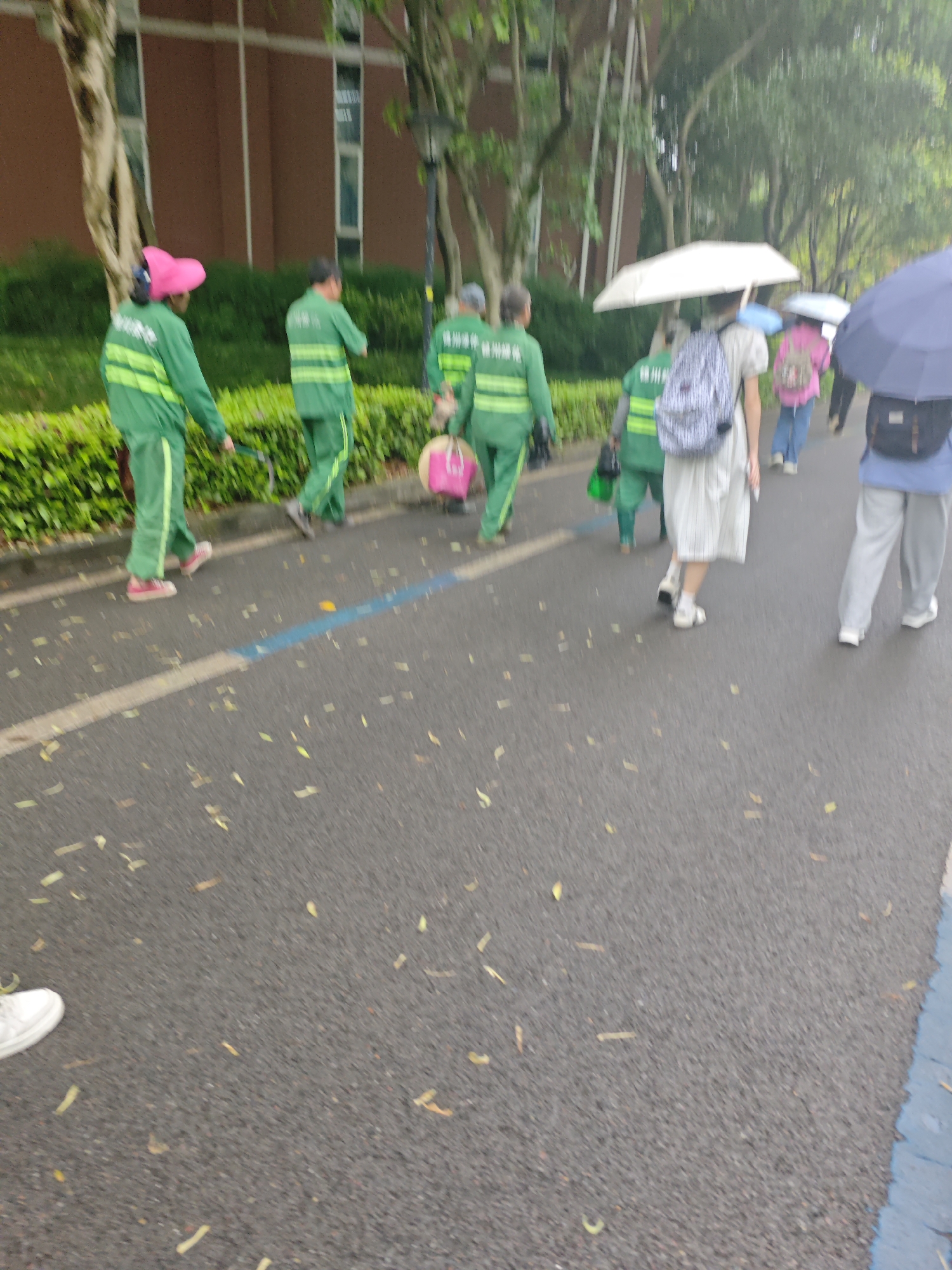 The image depicts a group of municipal sanitation workers in the Chongqing High-tech Zone, China, wearing green uniforms and walking along a paved road. One of the workers is wearing a pink hat. Beside them are other pedestrians, some carrying umbrellas, walking in the same direction. The road surface is wet and littered with fallen leaves. A building is visible in the background.