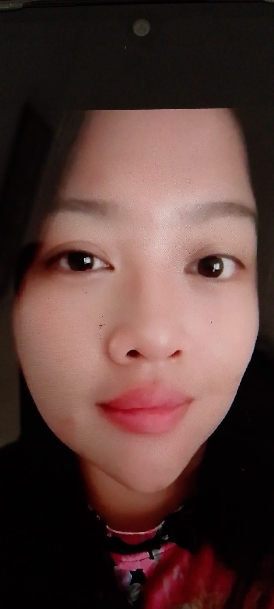 The image is a close-up, front-facing portrait of a young East Asian woman, likely taken with a mobile device. Her face fills most of the frame, with a portion of a dark device bezel visible at the top. She has dark brown eyes with a subtle sheen, neatly shaped eyebrows, and a small, delicate nose. Her lips are full and painted with a soft pink lipstick, and her skin appears smooth with a slight blush on her cheeks. The background is dark and out of focus, with a hint of a patterned garment with floral motifs in shades of pink and black visible around her neck. The overall lighting is soft, highlighting her features. The image appears to be a selfie, given the close framing and direct gaze.