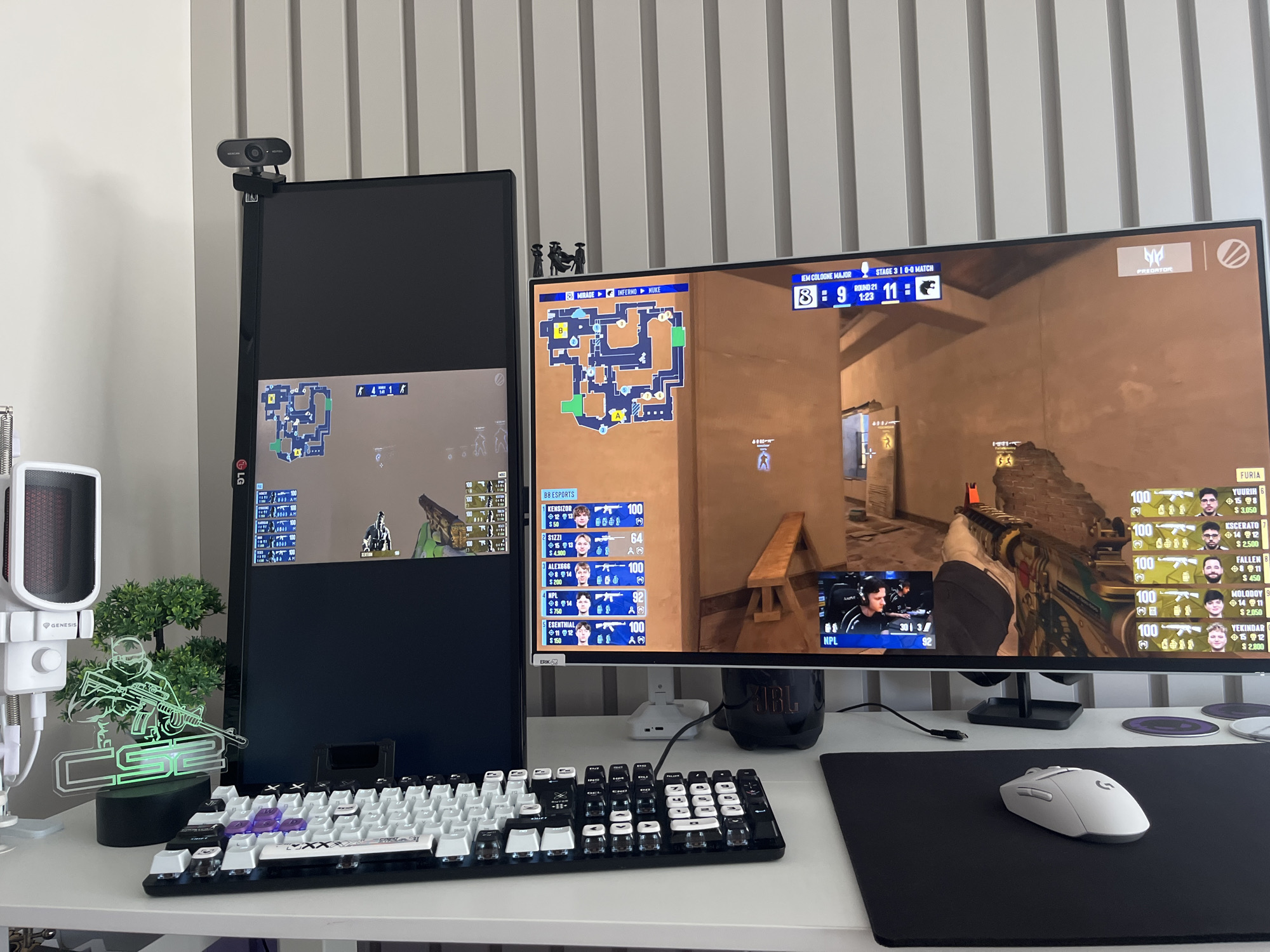 A gaming setup is displayed on a desk with a white wall in the background. Two monitors are present; a vertical monitor on the left displays a game map, and a horizontal monitor on the right shows gameplay with player statistics overlayed. A webcam is attached to the top of the vertical monitor. On the desk, a white and black keyboard is positioned in front of the monitors. To the left of the keyboard, a white microphone with a red grille sits on a stand. A small artificial tree and a green, glowing acrylic figurine of a gamer holding a rifle are placed between the microphone and the vertical monitor. A white wireless mouse rests on a black mousepad to the right of the keyboard. The environment is clean and organized, suggesting a dedicated gaming space. The scene does not provide any visual cues to determine its location in Varna, Bulgaria.