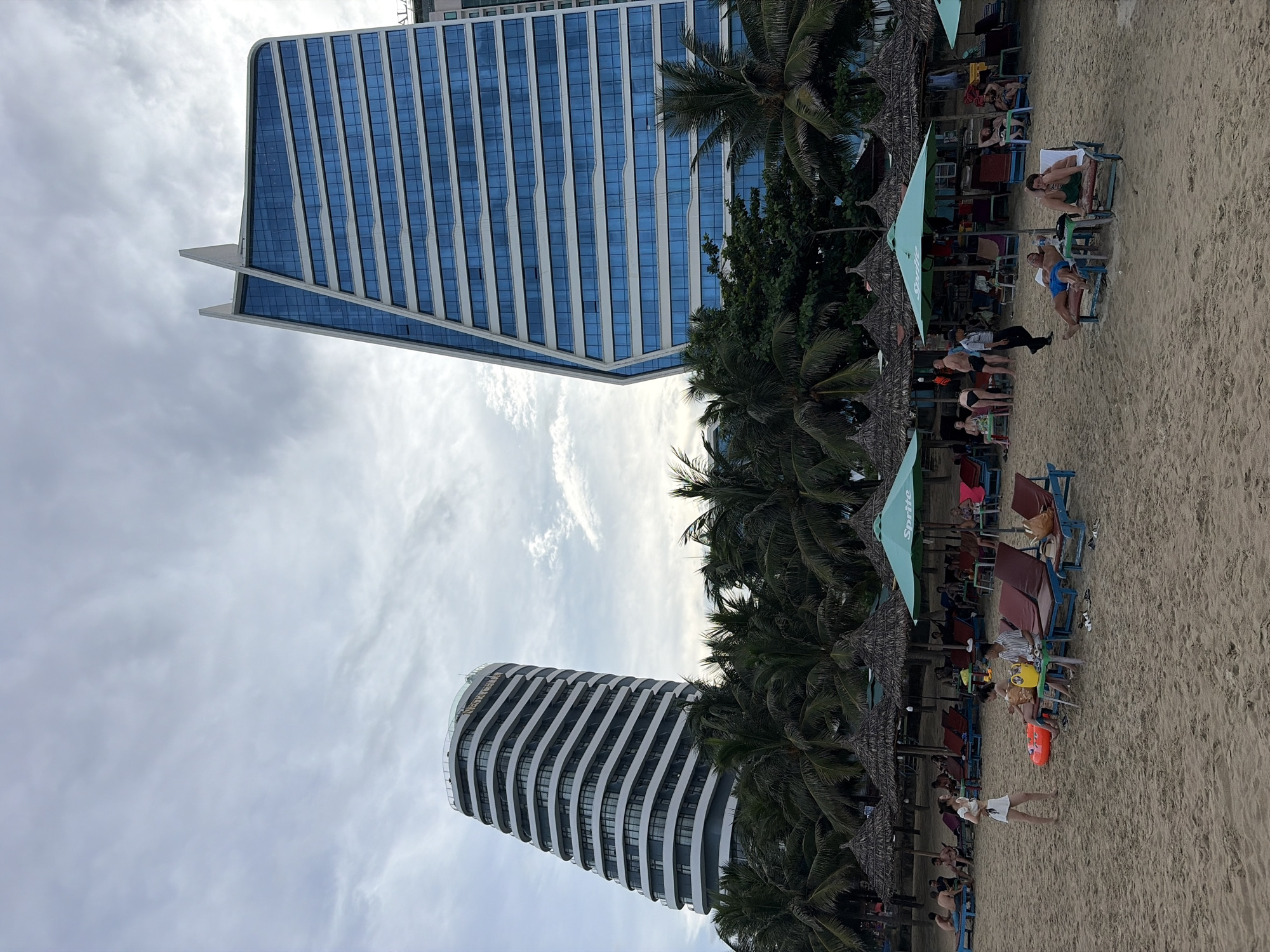 This is a photograph of a beach scene in Da Nang, Vietnam. Two modern, high-rise buildings dominate the background. The building on the left has a distinctive, wavy facade, while the building on the right is a sleek, angular structure with numerous blue-tinted windows. In front of the buildings, a line of palm trees separates the beach from the structures. The beach itself is covered in sand, with many beach chairs and umbrellas set up. Several people are visible, some lounging on chairs, others walking on the sand. The sky is overcast with some scattered clouds. The overall atmosphere suggests a public beach area in a city with modern architecture.