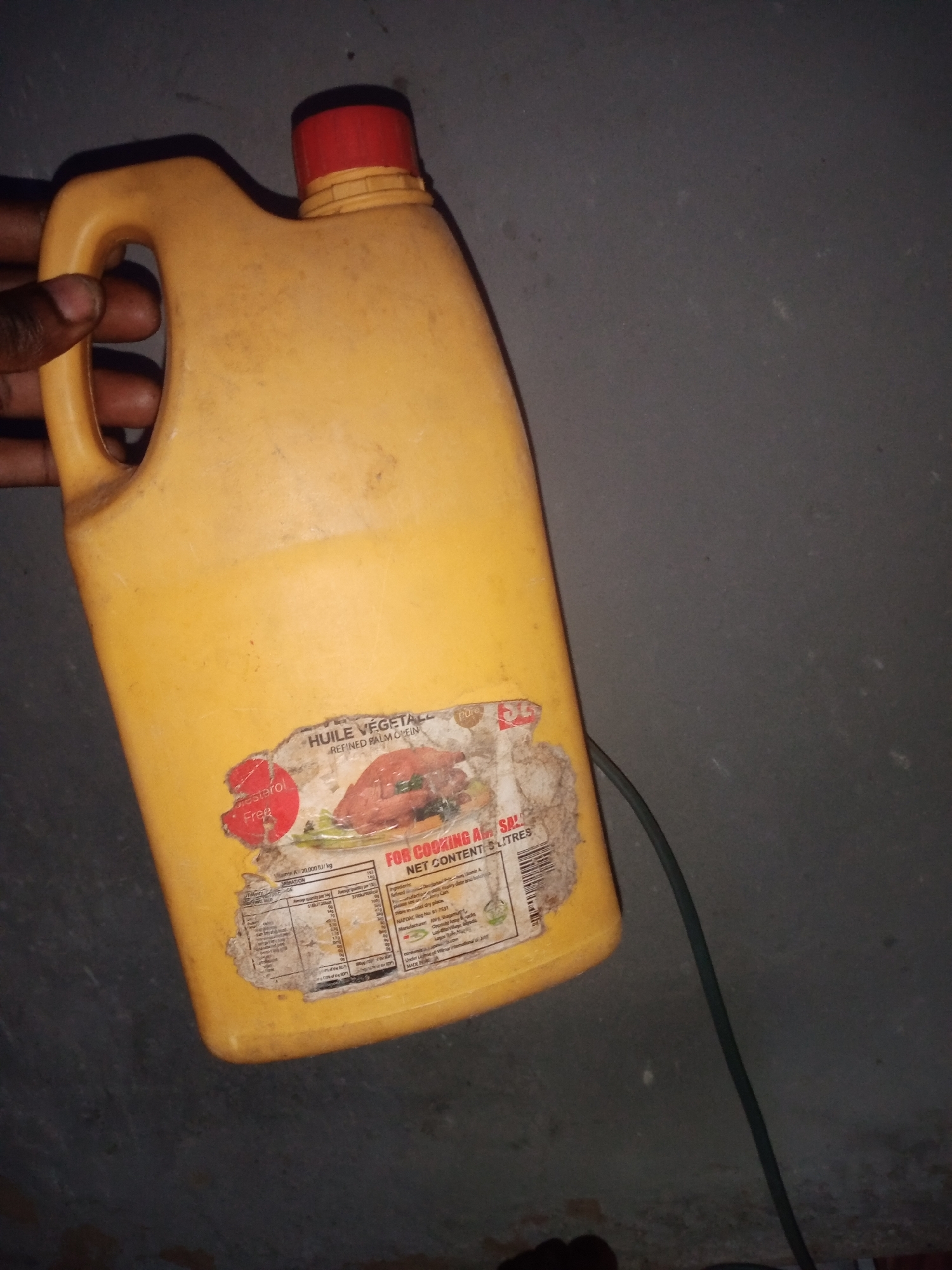 A person's hand is holding the handle of a large, yellow plastic jug of cooking oil. The jug has a red cap. A label on the jug is partially torn and smudged, but it is possible to read some text, including "HUILE VEGETALE," "REFINED PALM OLEIN," and "FOR COOKING." Nutritional information and other text are also present on the label. The jug is placed against a dark, textured wall, possibly concrete. A thin, dark green cord or tube is visible to the right of the jug. The lighting is dim, casting shadows on the wall. The scene appears to be indoors or in a sheltered outdoor space, with no clear indicators of a specific city location other than the user's prompt.