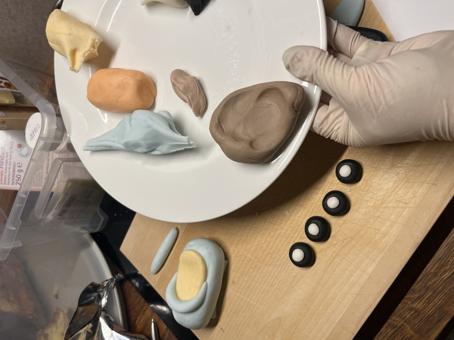 A white plate holds lumps of yellow, orange, light blue, two shades of brown, and black fondant. A gloved hand holds the plate over a wooden cutting board. Four black fondant circles, each with a smaller white fondant circle in the center, sit on the board. Below them is a blue fondant shape with a yellow fondant piece attached and a small blue fondant roll. The items appear to be for decorating or making a cake.