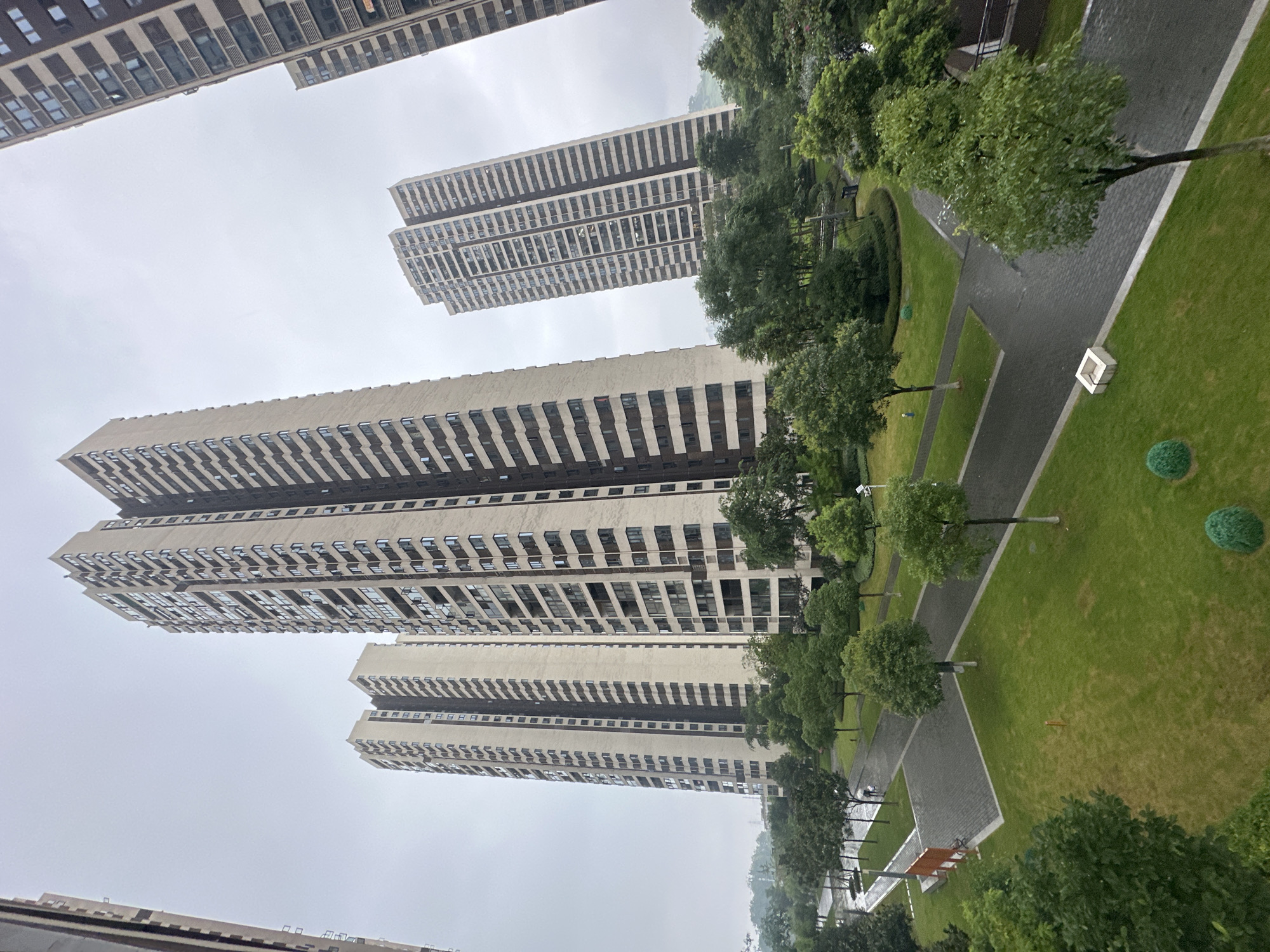 The image depicts a residential complex in the Chongqing High-tech Zone, China. Several tall, modern apartment buildings with beige and dark grey facades dominate the background. The buildings feature numerous windows and balconies. In the foreground, there is a well-maintained park area with lush green lawns, neatly trimmed trees, and paved pathways. Two individuals are walking on one of the pathways. The sky is overcast, suggesting a cloudy or possibly rainy day, and the overall atmosphere is calm and orderly.