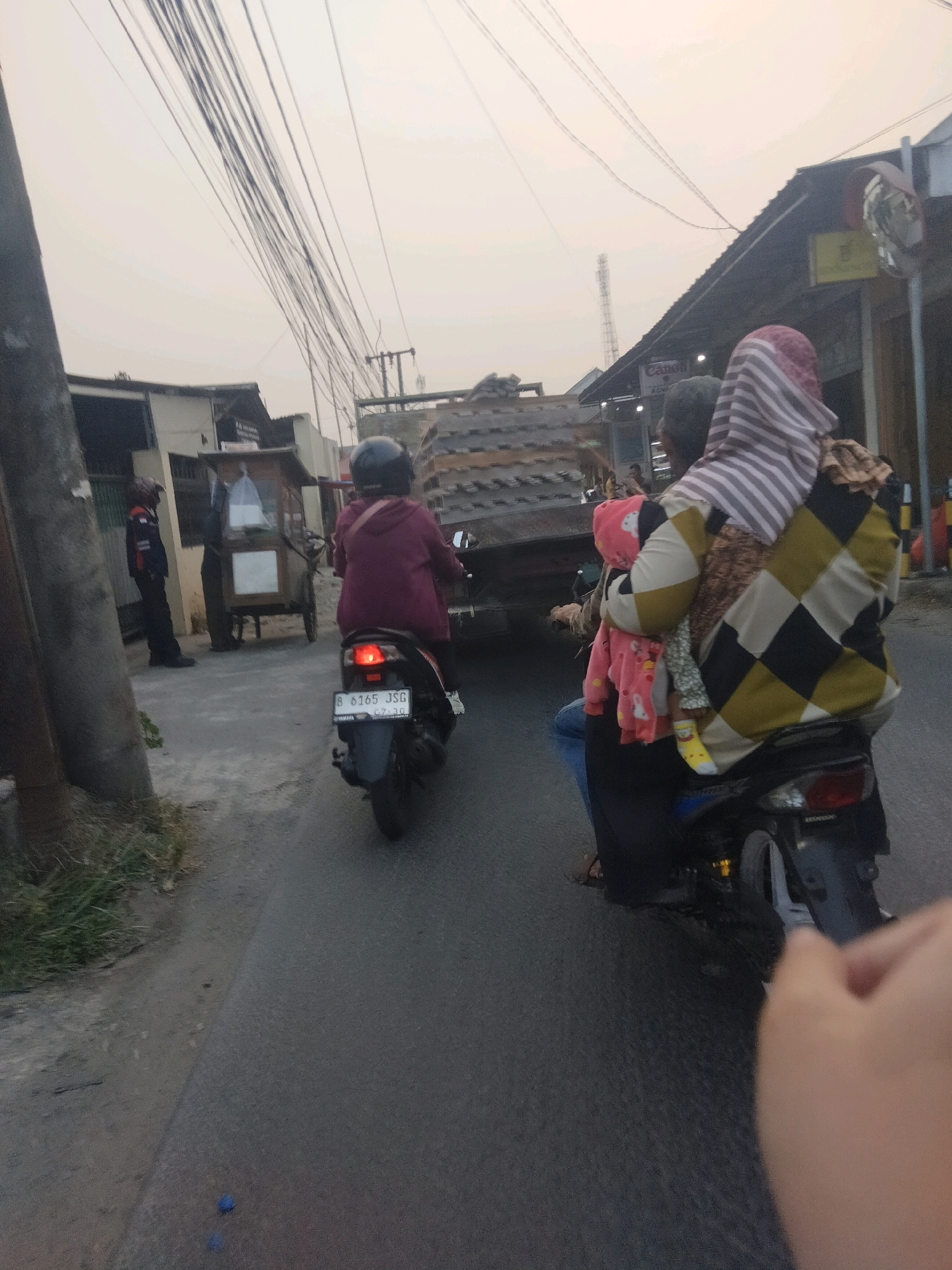 The image shows a street scene in Pisangan Jaya, Indonesia, with a focus on two motorcycles on a paved road. The sky is overcast, and a tangle of overhead power lines dominates the upper portion of the image. On the left, a motorcycle with a rider wearing a maroon jacket and a black helmet is moving away from the camera. The motorcycle's license plate reads "B 6165 JSG". To the right of this motorcycle, on the sidewalk area, a person in uniform with a red stripe on their sleeve and black pants is standing next to a wheeled cart.
In the foreground and to the right, another motorcycle is seen from the rear. This motorcycle carries two passengers and a child. The adult passenger in the back is wearing a patterned headscarf and a jacket with a diamond pattern in yellow, black, and beige. They are holding a young child who is wearing a pink jacket with a white teddy bear print. The child is also wearing a yellow sock on their right foot. The passenger in front appears to be wearing blue jeans.
In the background, there are buildings, one with a dark, sloped roof and another structure with wooden siding. A large truck with a load of what appears to be metal scaffolding or corrugated panels is positioned further down the road. The road surface is asphalt, and there is some dry grass and dirt on the left side. The overall impression is of a typical street scene with various modes of transportation and urban infrastructure.
