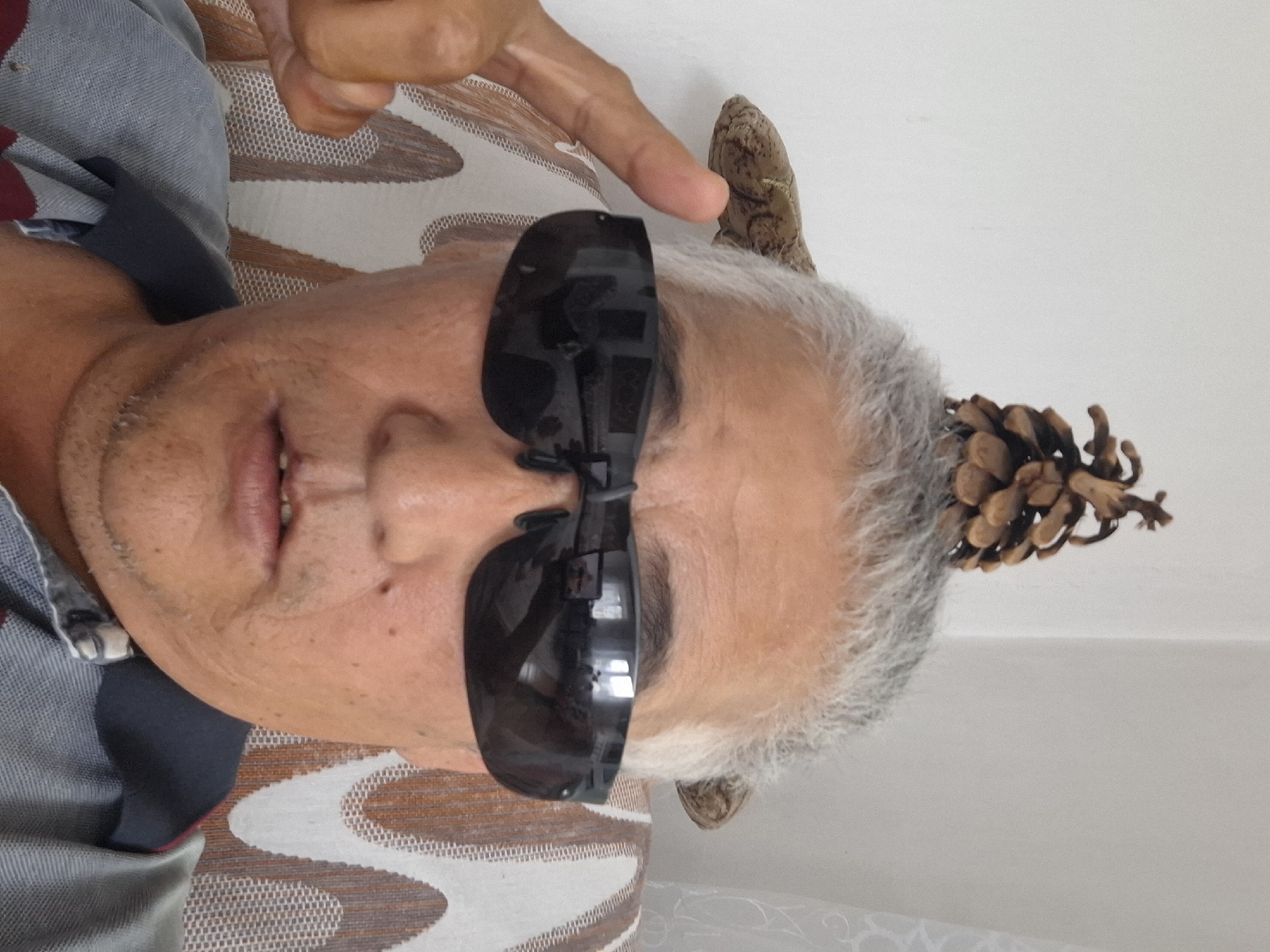 A man with gray hair and sunglasses is sitting in a chair. A pine cone is balanced on top of his head, and a piece of wood is behind his ear. The man is looking at the camera. The background is a white wall with a patterned chair. The image appears to be taken indoors. There is no identifiable information in the image or accompanying text to indicate a specific location within the city of Almaty, Kazakhstan.