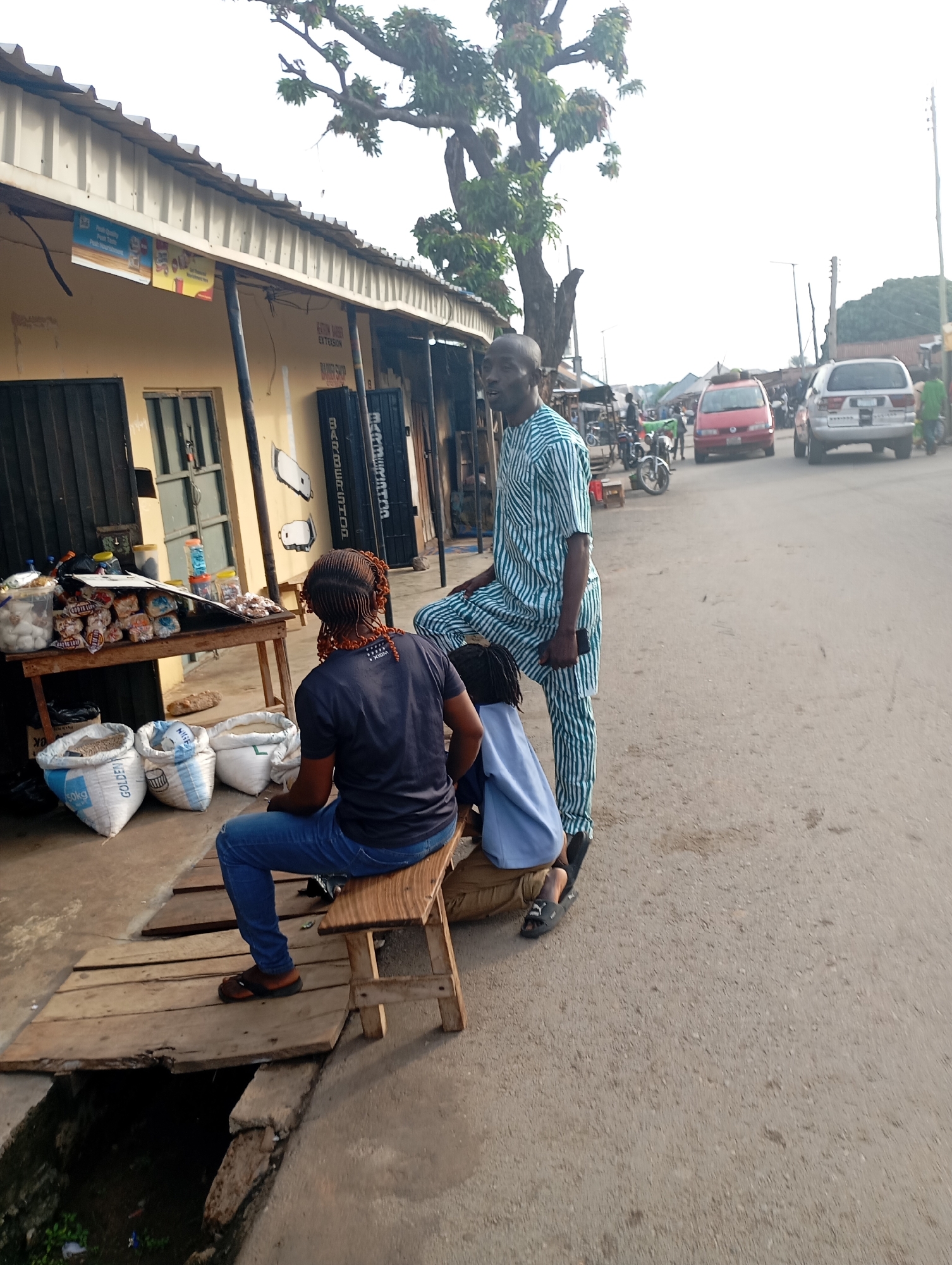 A medium shot shows a street scene in Shendam, Nigeria. In the foreground, a woman with braided hair sits on a wooden bench, facing away from the camera. She is wearing a dark t-shirt and blue jeans. Next to her, a man in a green and white striped traditional outfit stands, holding a black mobile phone in his right hand. He is looking to the left. To the left of the bench are three large sacks of grain. Behind the bench, a wooden table displays various goods, including bags of bread, plastic bottles, and jars. The woman's legs and feet are visible, as she is sitting on a wooden bench that is placed over a drainage ditch.
In the background, a street with vehicles is visible. A red van and a silver SUV are driving down the road. Several motorcycles are parked on the side of the road. Buildings line the street, with awnings and signage. A large tree with leafy branches extends over the scene. The sky is overcast.