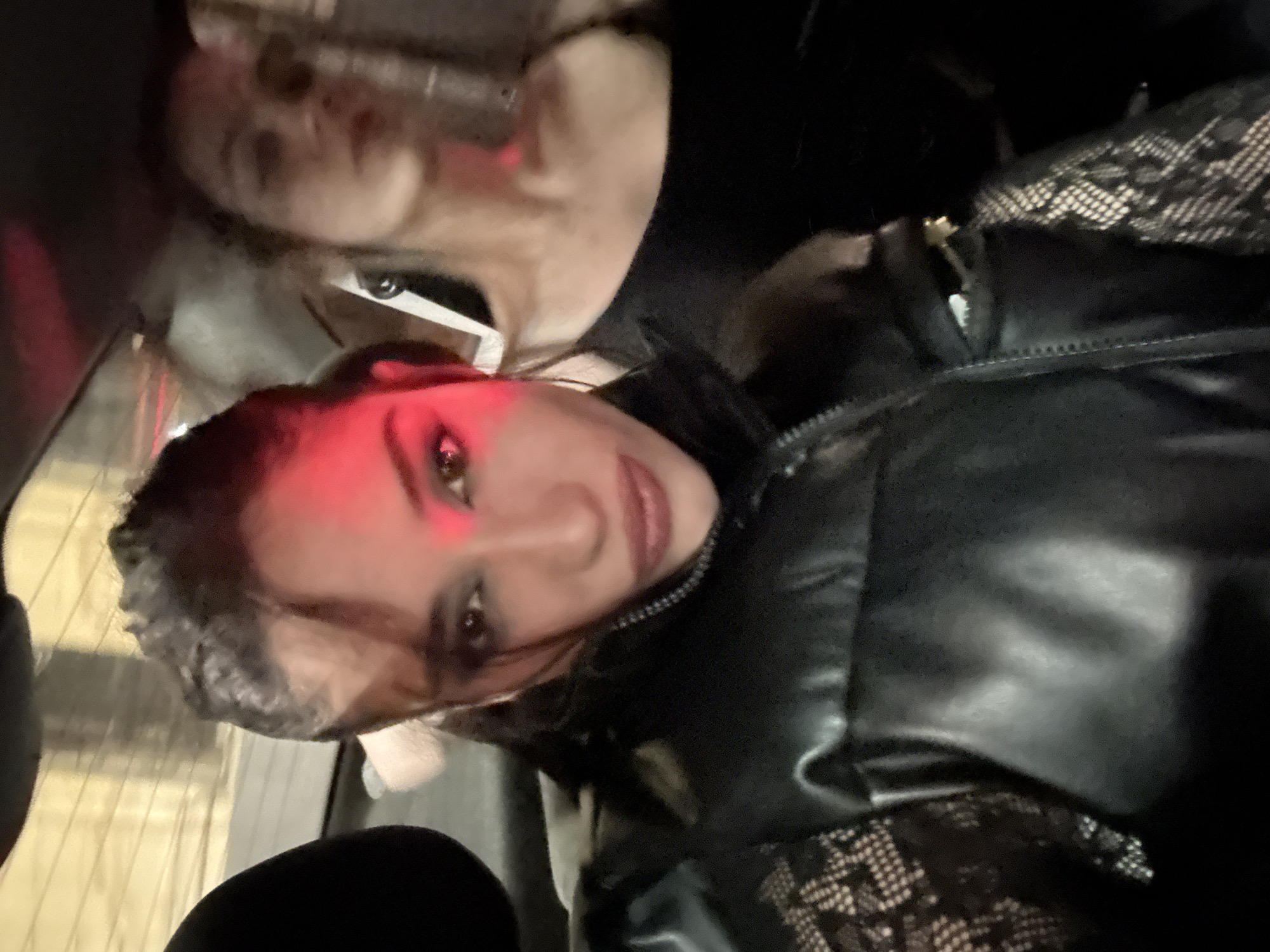 The image shows two women indoors, possibly in a car. The foreground woman is wearing a black leather jacket over a lacy black top. Her makeup includes dark eyeshadow and a red light is reflecting off her face. Behind her is another woman wearing sunglasses and a black top. A car window and seat are visible. It's impossible to pinpoint a specific location in Buenos Aires based solely on this image.